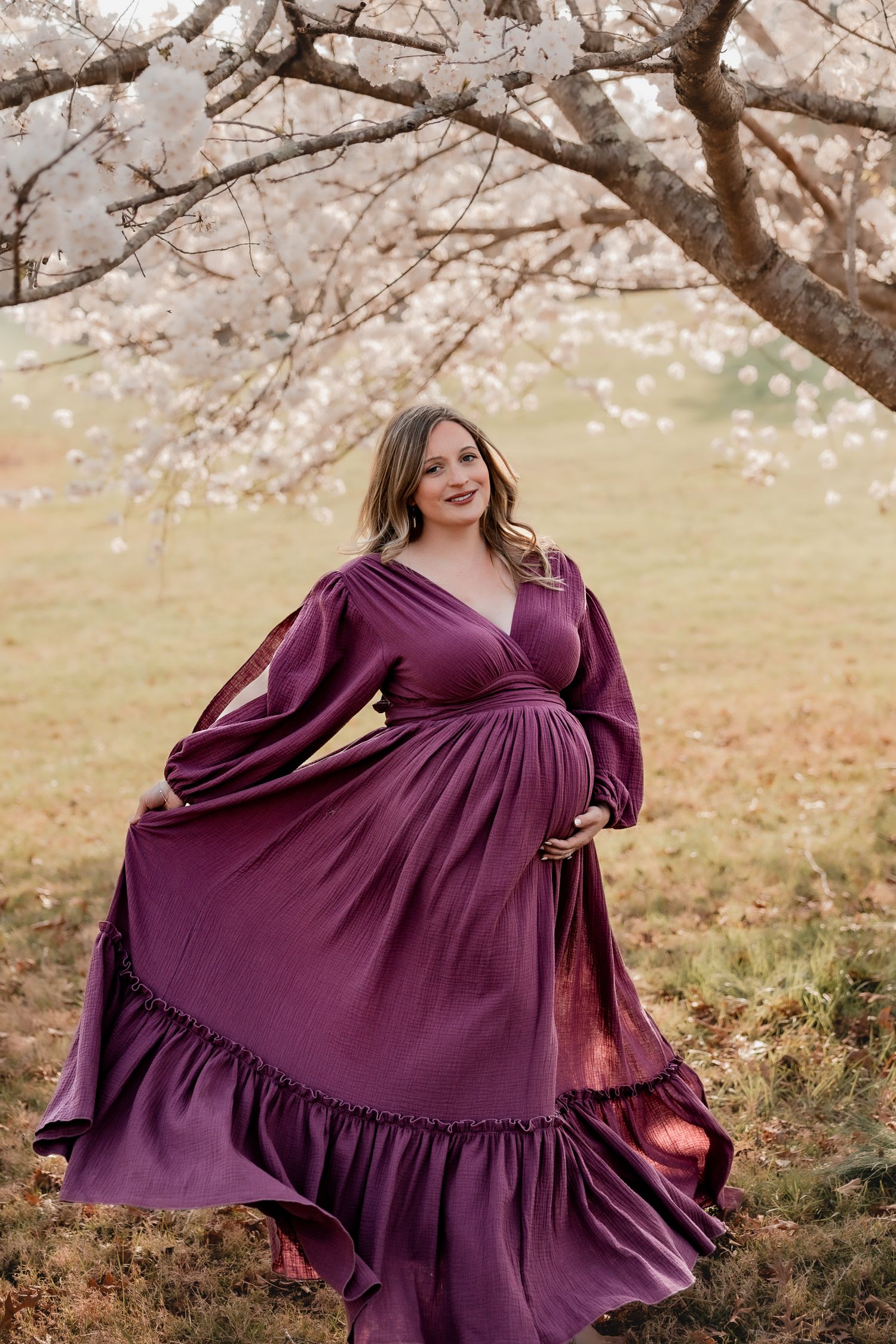 Pregnant woman in flowing purple maternity gown poses under cherry blossom trees in a serene outdoor setting.
