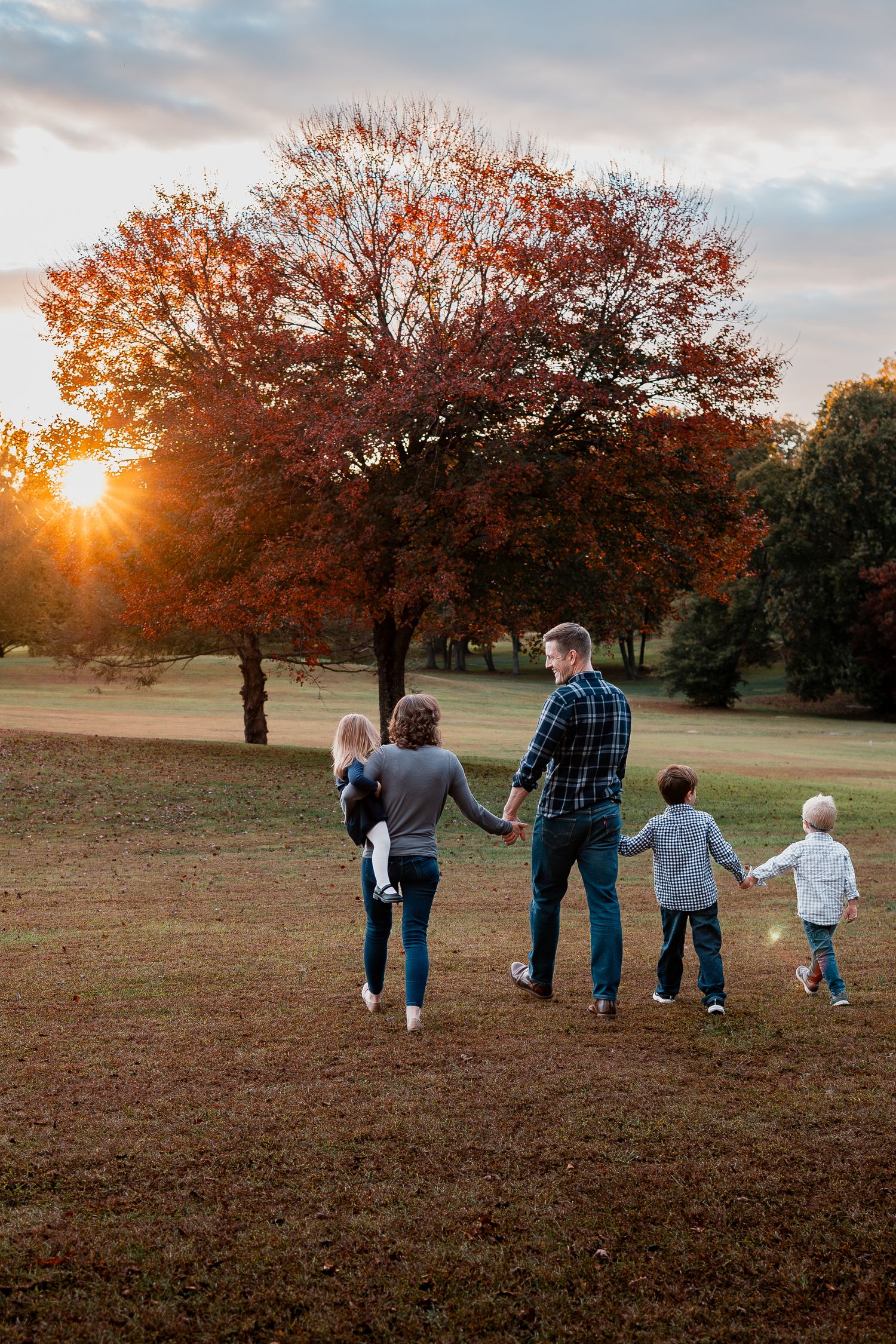 A family walks hand in hand across a grassy field at sunset with autumn trees in the background.