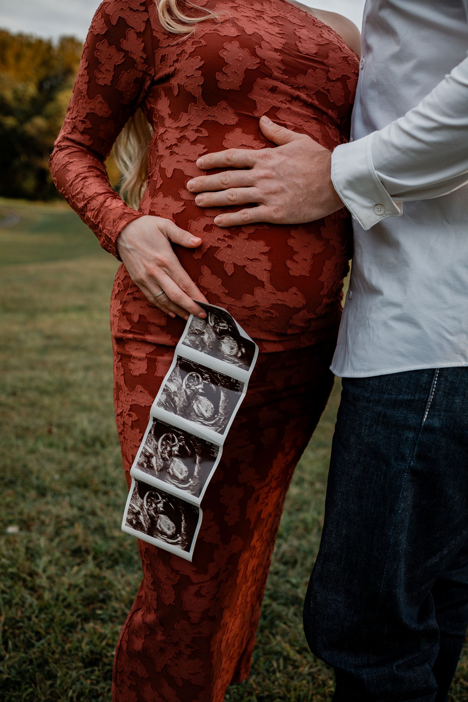 A couple in a red dress and white shirt hold ultrasound photos while standing in a grassy field.