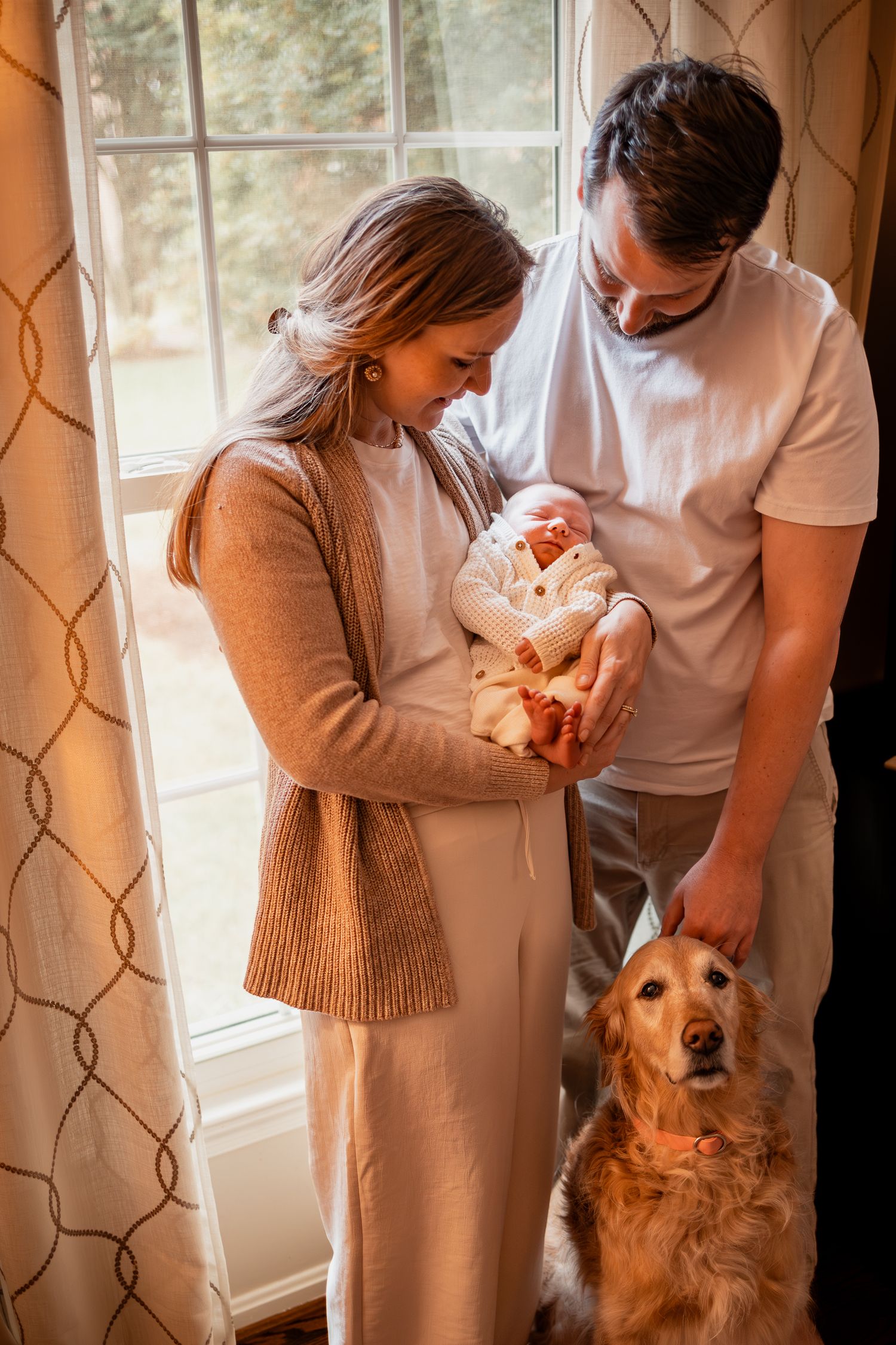 A couple stands by a window with their golden retriever dog while holding a newborn baby in warm natural light.