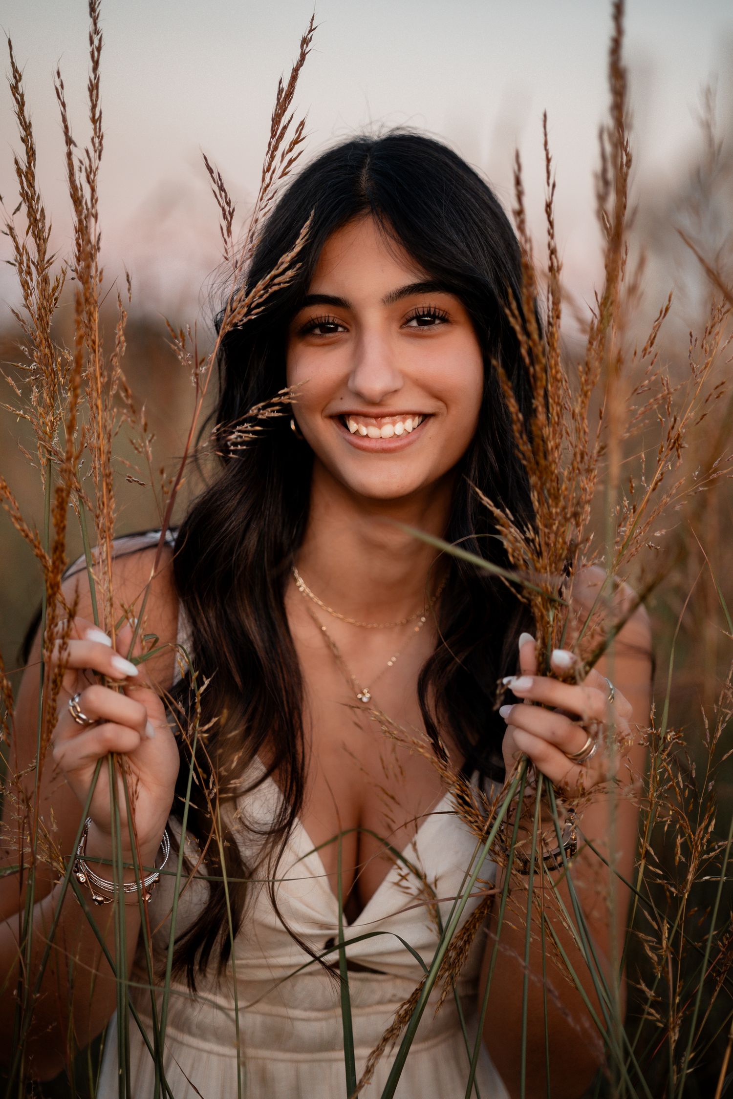Woman with dark hair poses in a grassy field wearing a white top during golden hour lighting.
