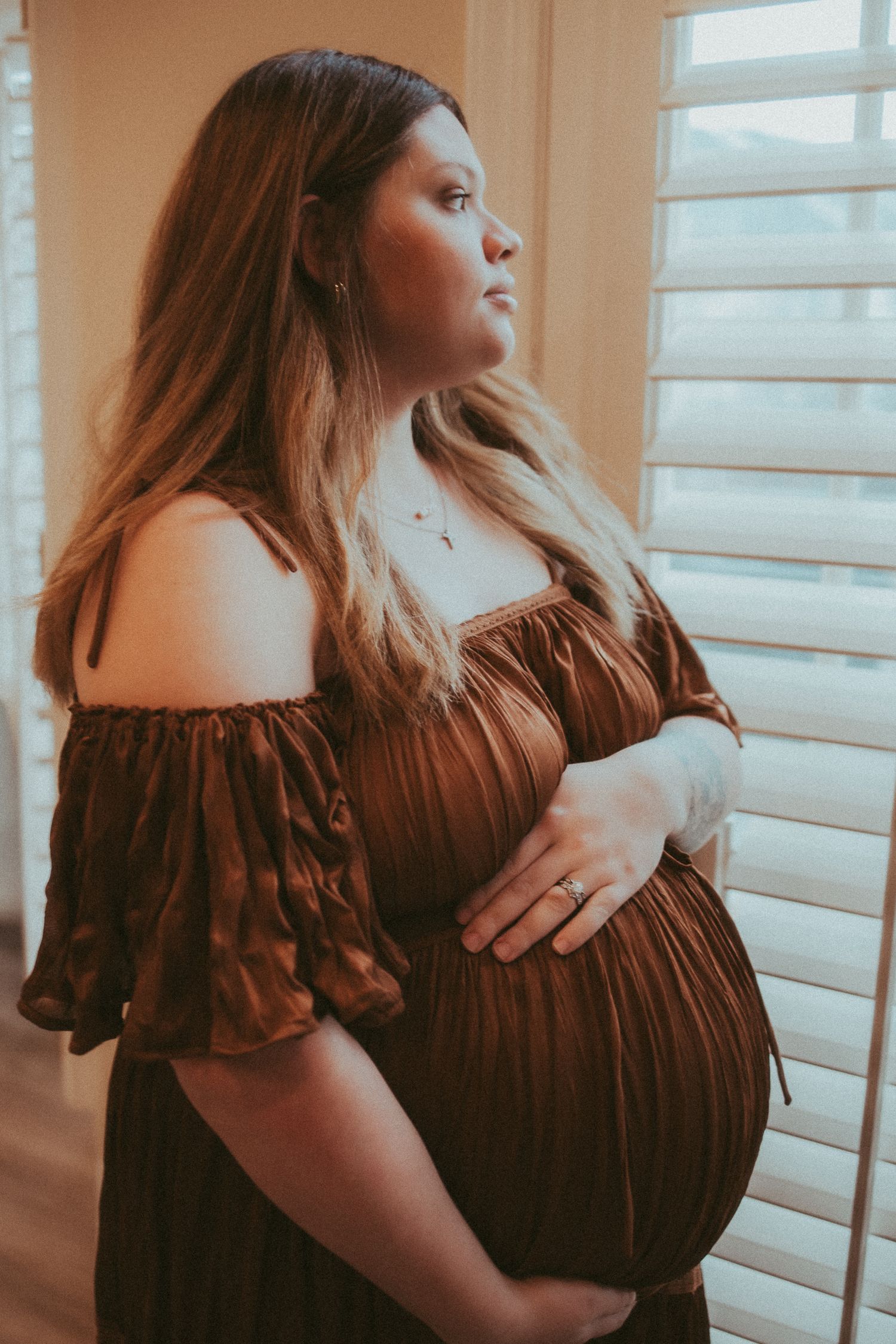 A pregnant woman in a brown off-shoulder dress stands by a window in soft natural lighting.