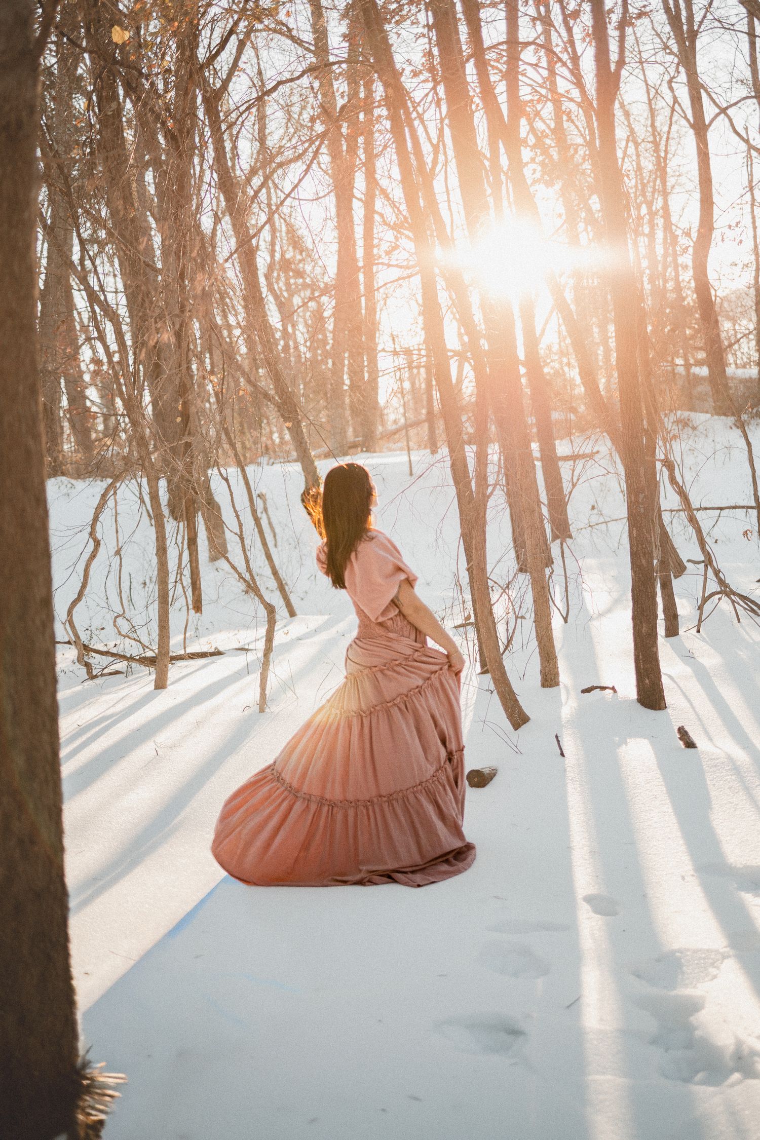 A person in a flowing pink vintage dress stands in a snowy forest at sunset with sunlight streaming through bare trees.