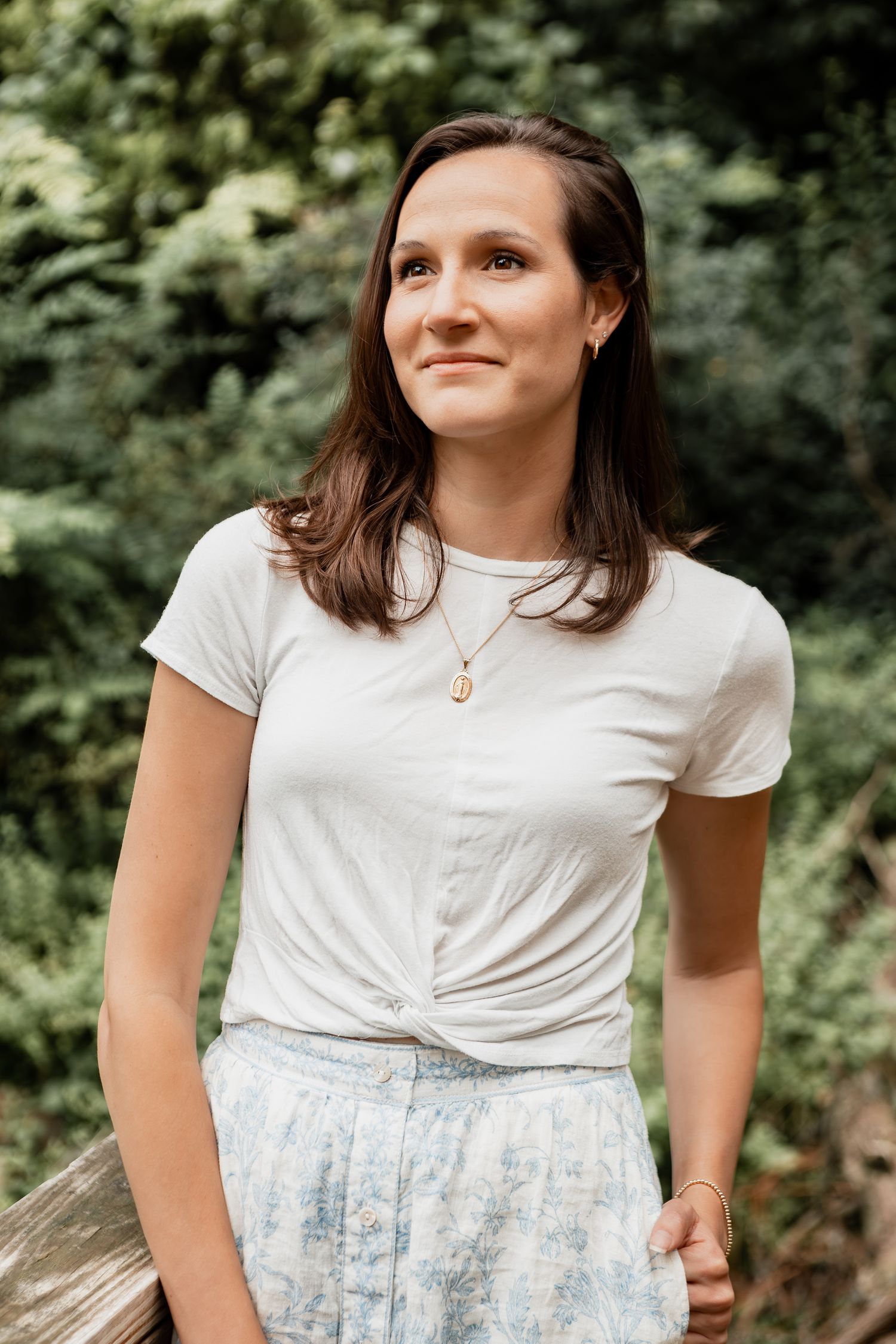 Portrait shot of woman in casual white t-shirt and patterned skirt against green garden backdrop.
