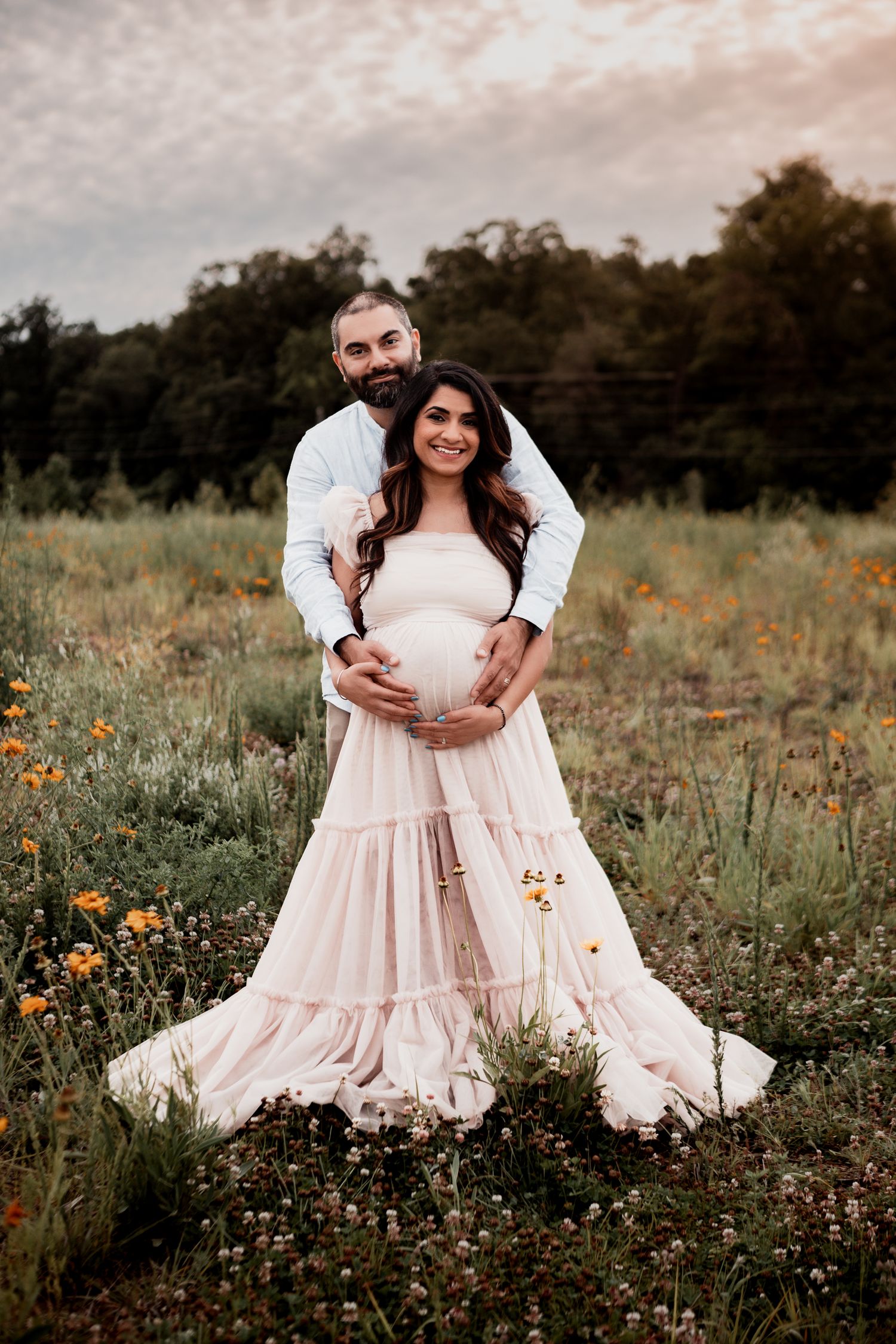 A couple poses together in a wildflower meadow at sunset, wearing soft flowy attire surrounded by yellow blooms.