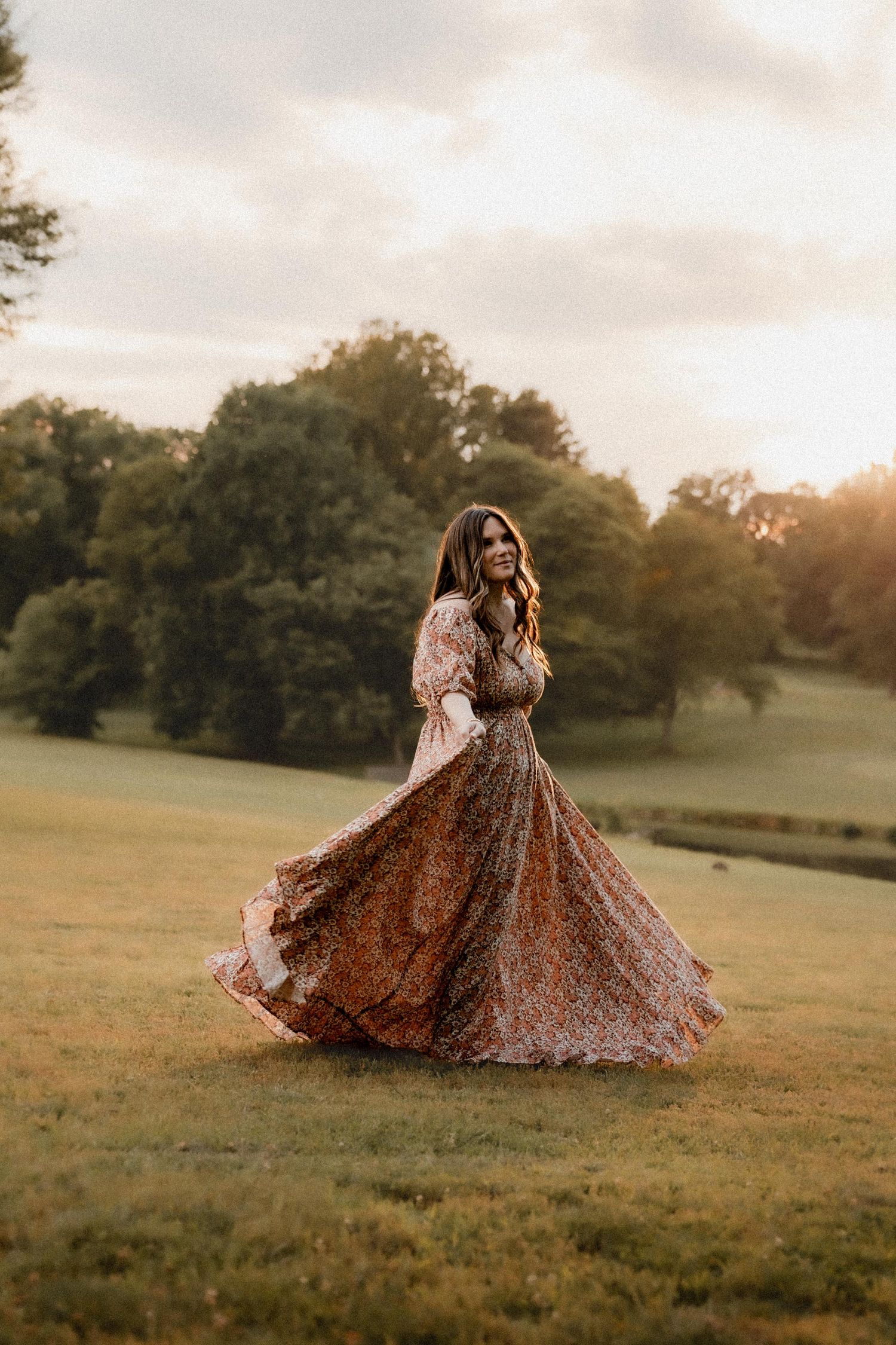 Woman in floral maxi dress twirling in golden hour sunlight on lush green field with trees in background.