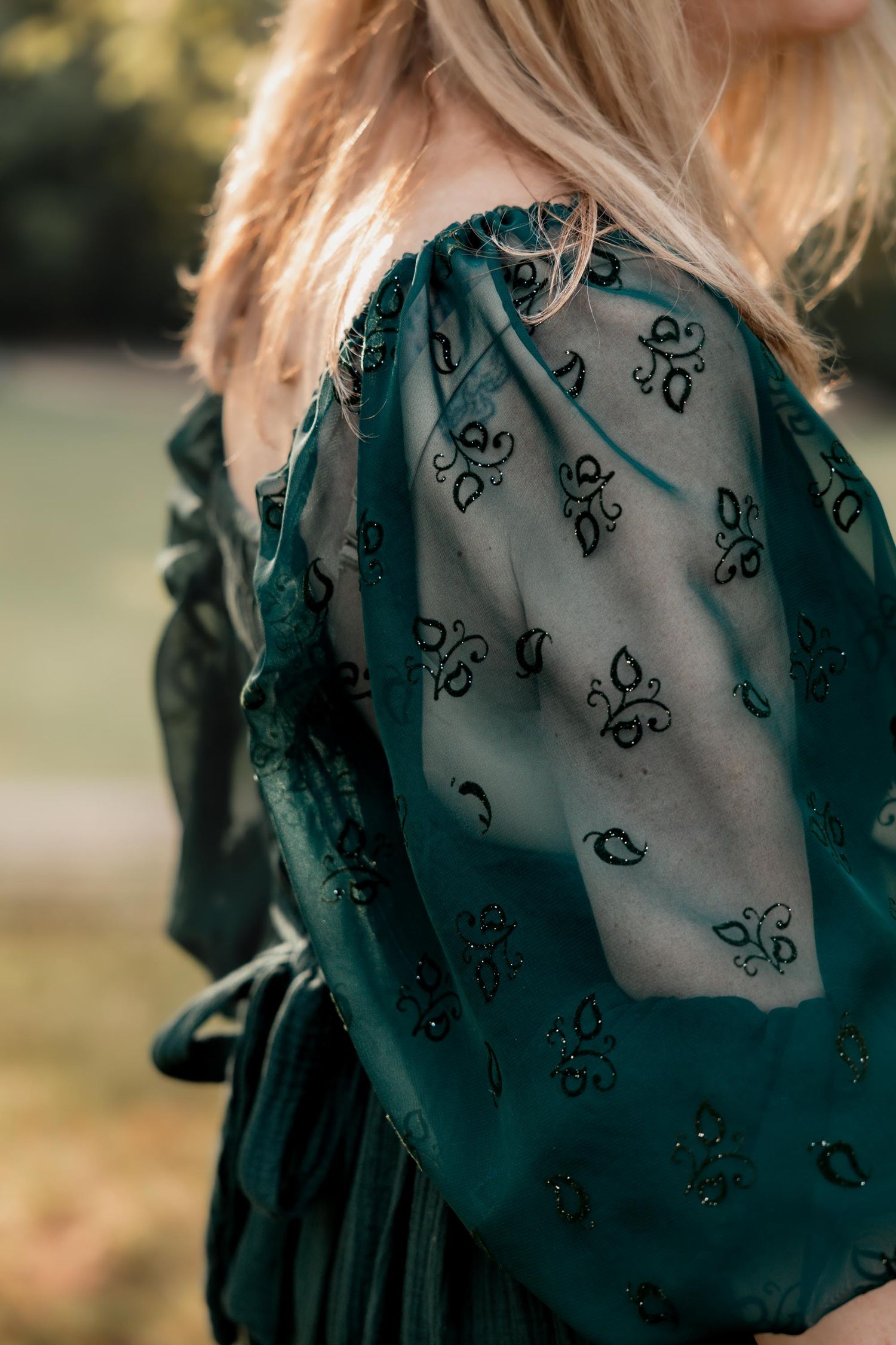 Close-up of a teal floral embroidered dress with sheer sleeves worn by a blonde woman outdoors.