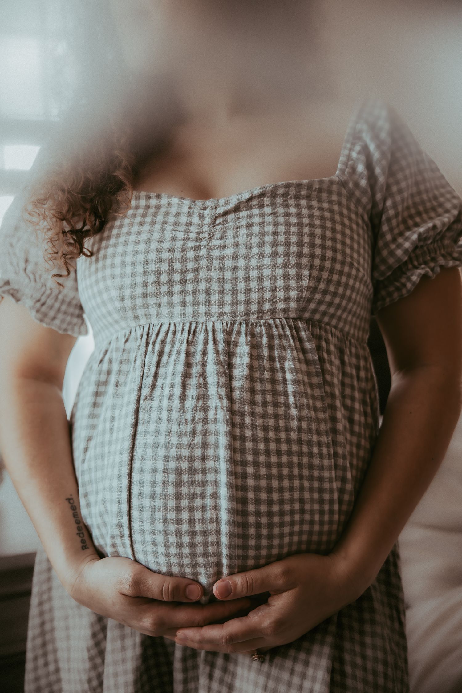 Pregnant woman in gingham dress cradling baby bump with both hands, soft natural light maternity photo.