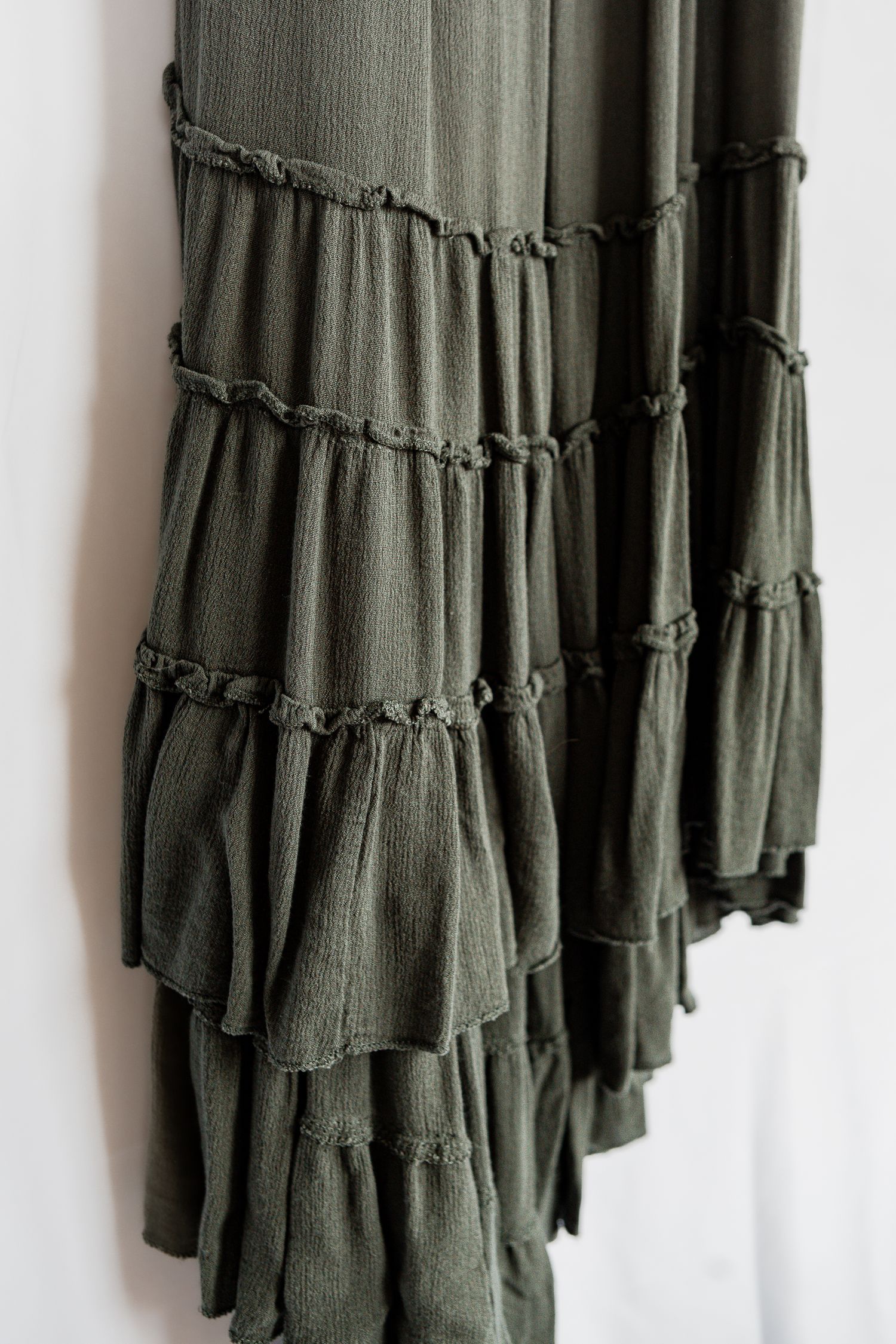 Close-up of olive green ruffled maxi skirt with tiered layers and gathered details hanging against white wall.