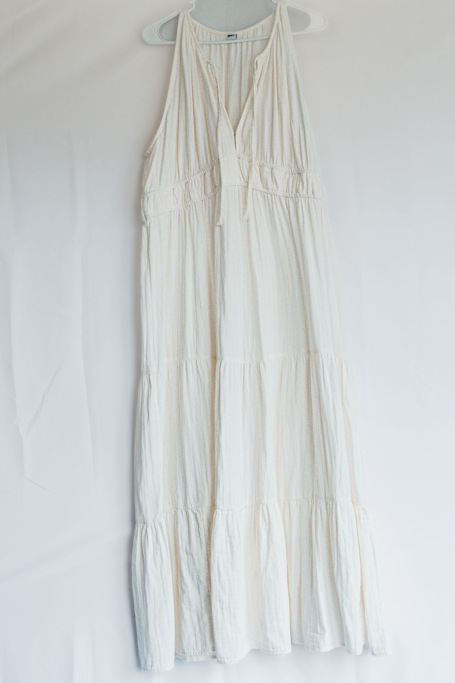 A white sleeveless tiered maxi dress with v-neckline and gathered waist detail hangs against a white background.