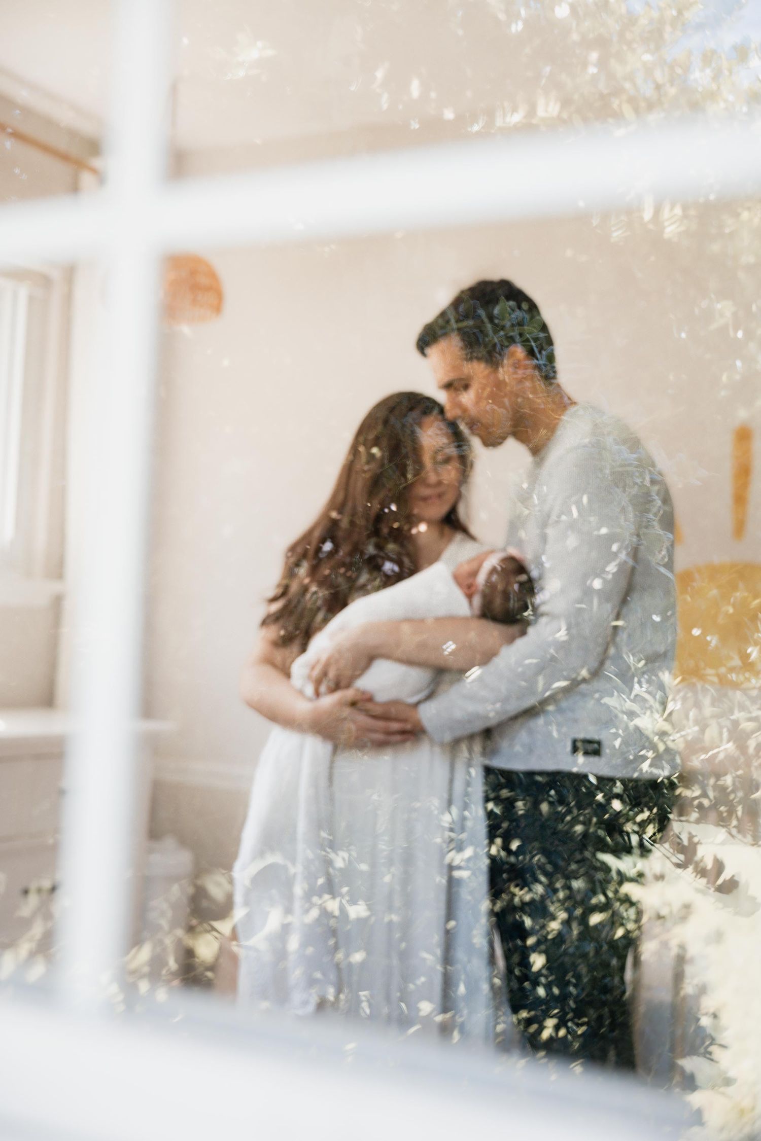 A couple tenderly holds their newborn baby, photographed through a window with soft natural light.