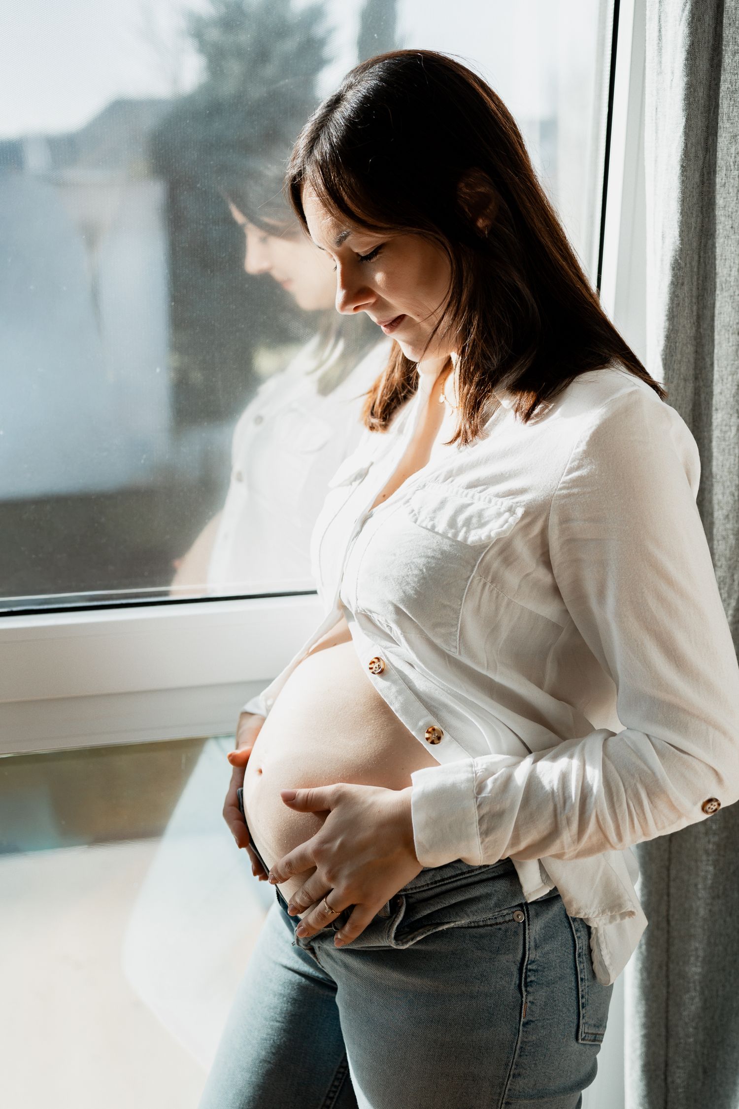 A pregnant woman in white shirt and jeans stands by window, gently cradling her growing baby bump.