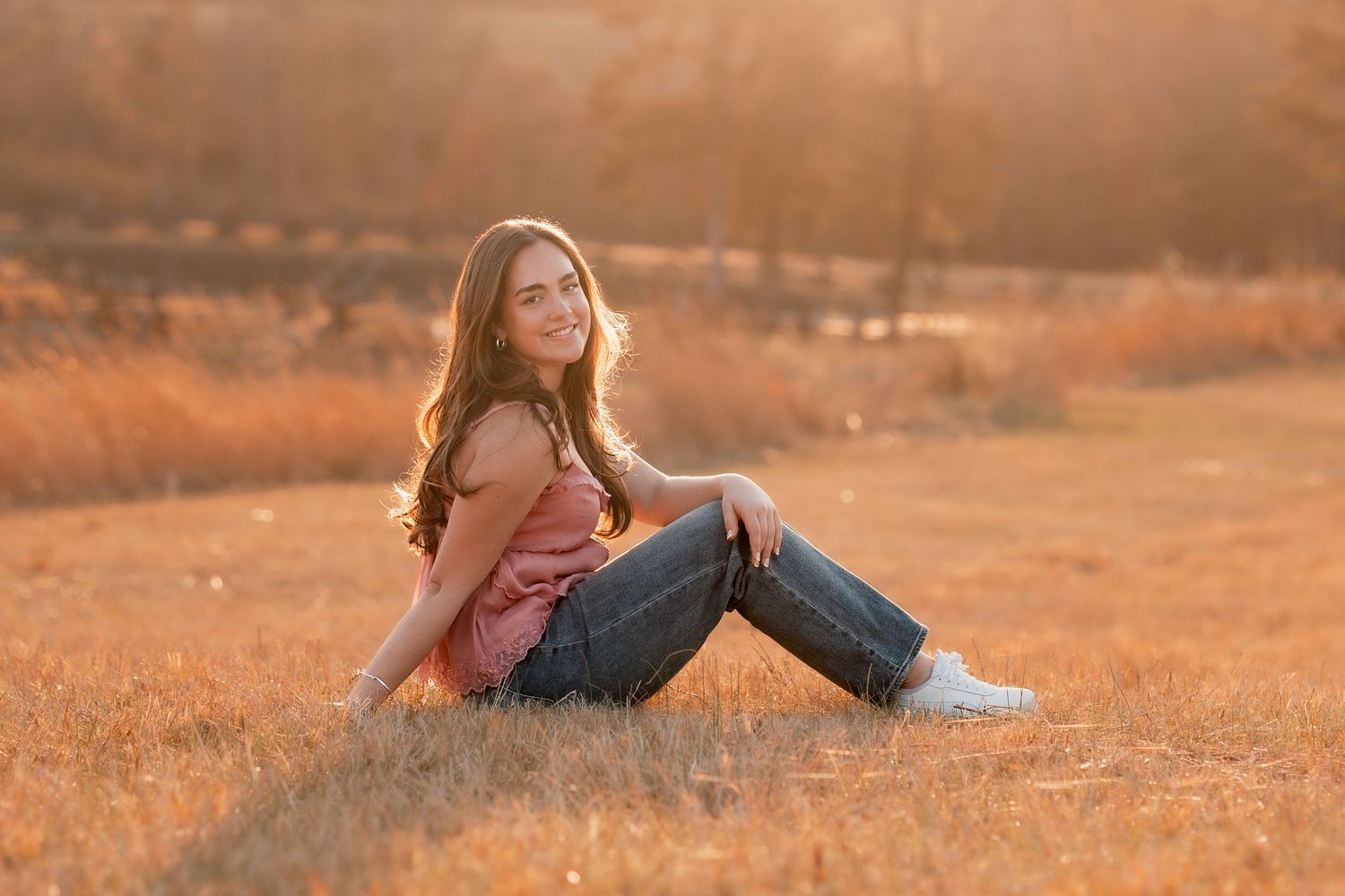 Young woman with long hair sitting in golden field at sunset, smiling, wearing pink top and jeans.