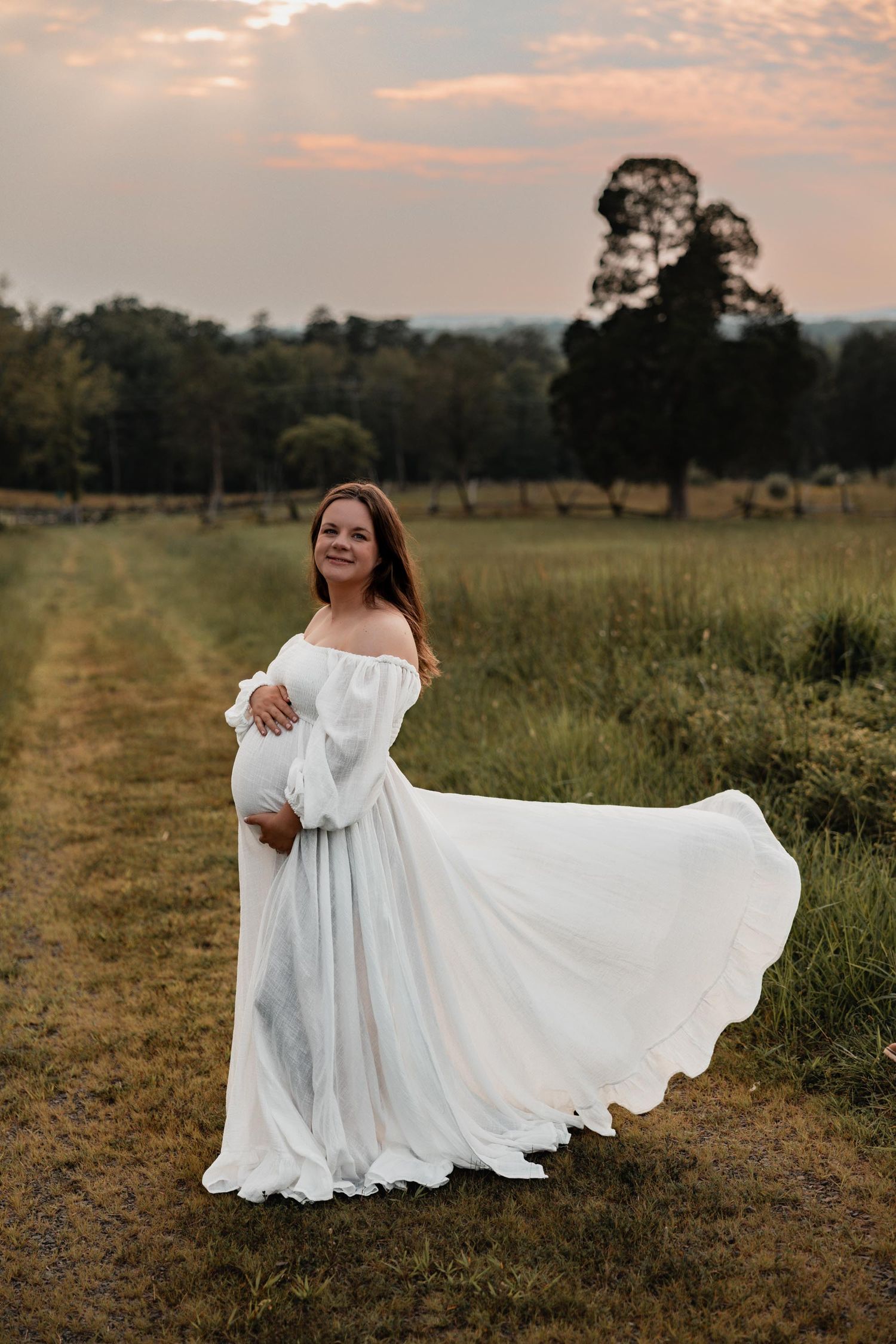 Radiant pregnant woman in billowing white off-shoulder dress posing on countryside path at golden hour sunset.