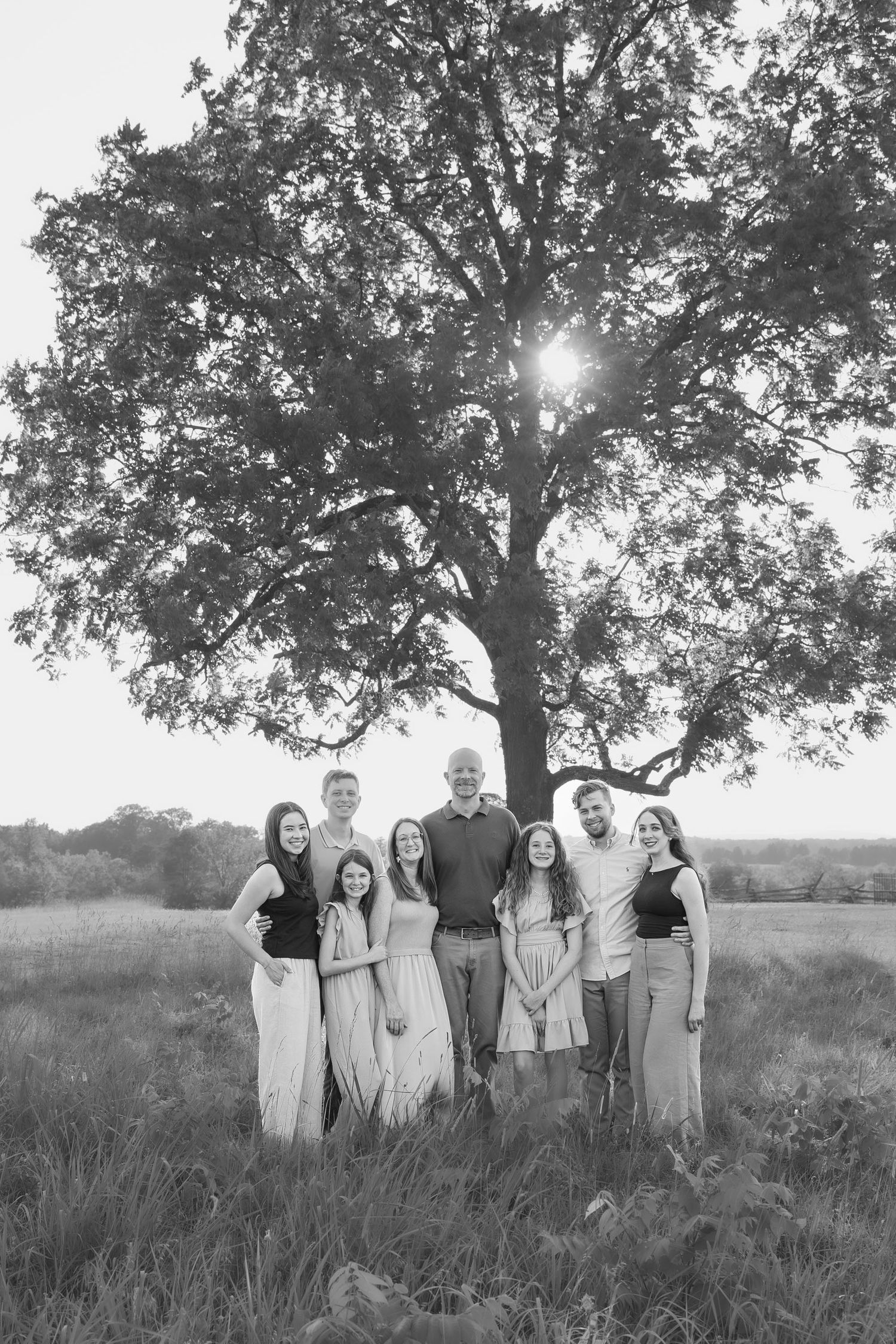 Black and white family portrait of seven people standing under a large oak tree in an open field.
