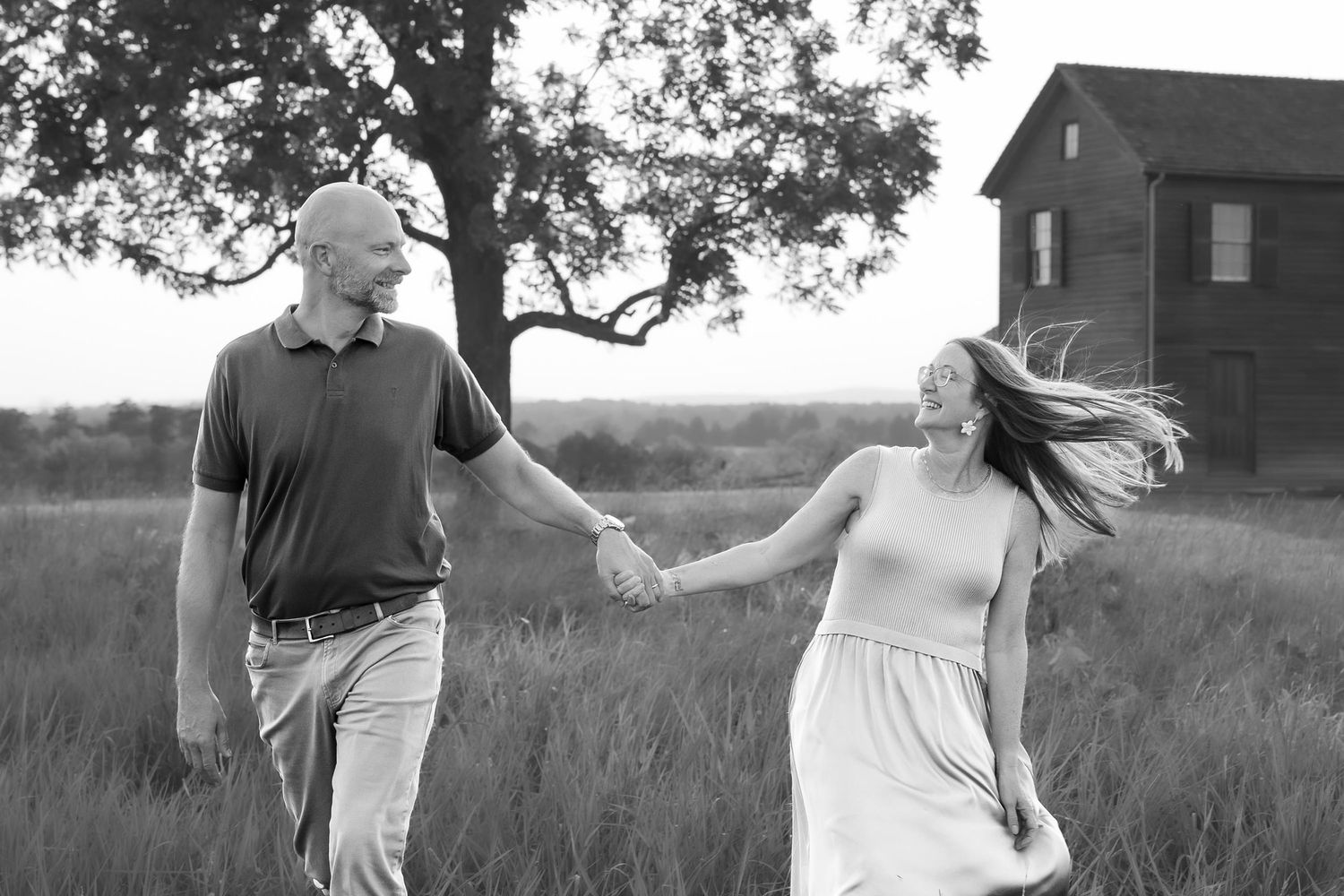 Black and white photo of a couple holding hands and walking through a field near a rustic barn and tree.