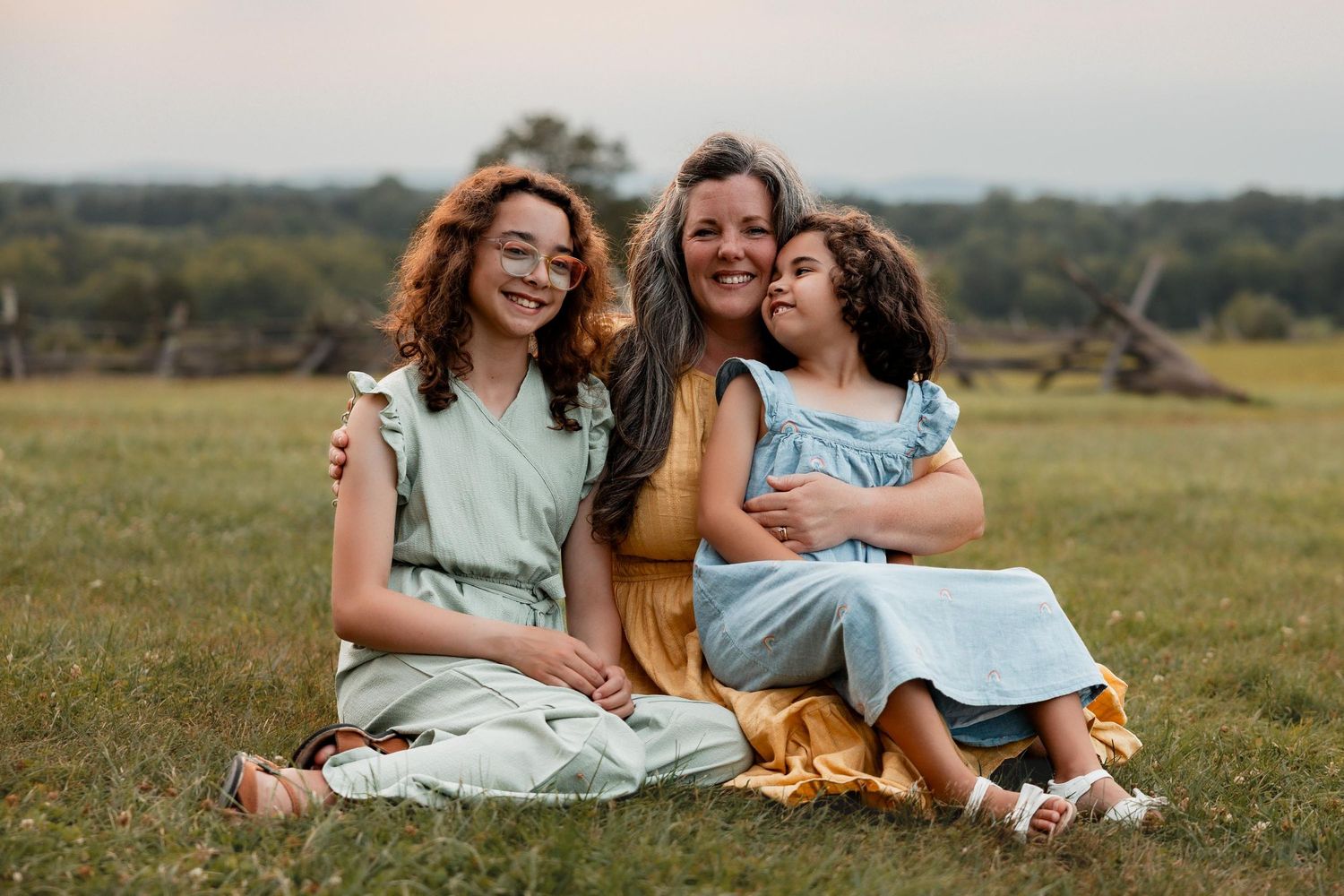 A mother sits in a grassy field smiling with her two young daughters during an outdoor family portrait session.