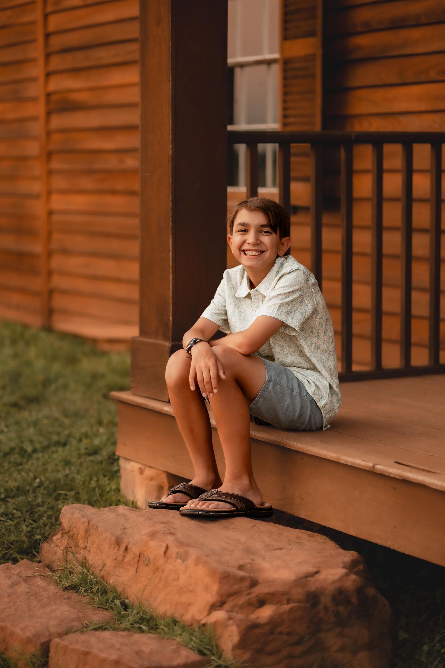 A smiling young boy sits on wooden porch steps in a floral shirt and denim shorts, wearing sandals.