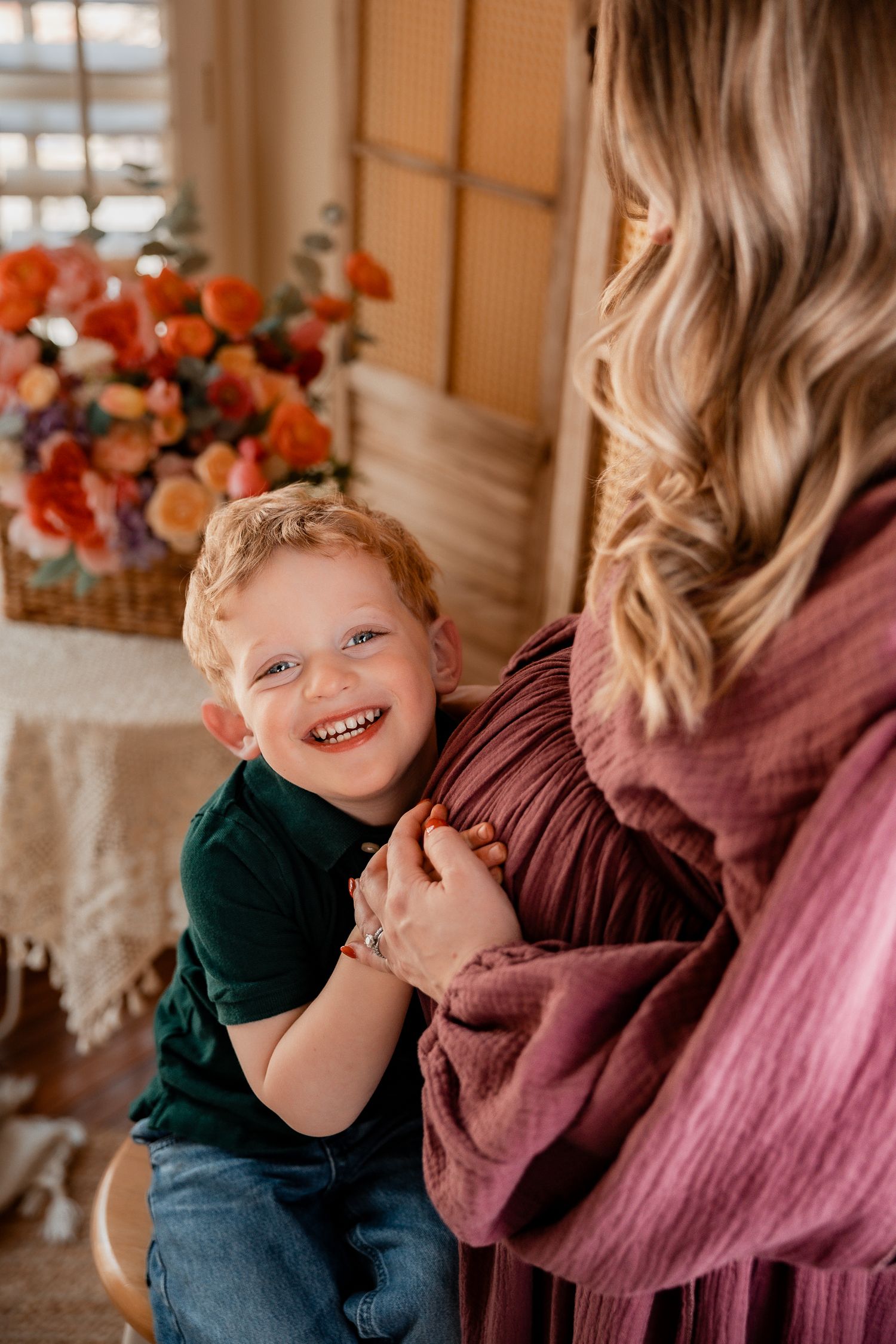 Happy red-haired toddler boy smiling while hugging pregnant mothers baby bump in floral decorated room.