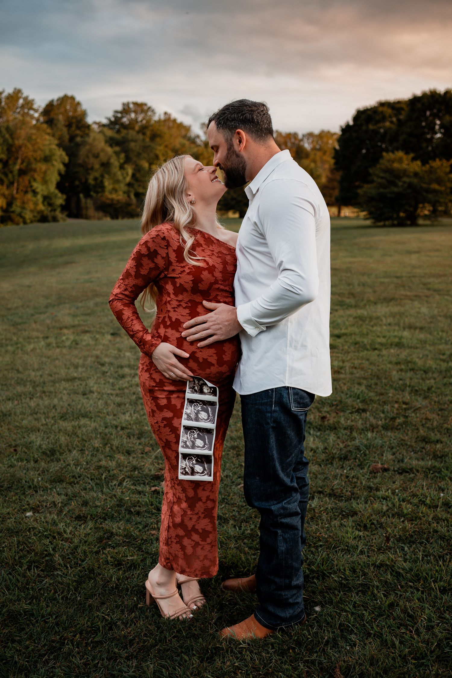 A couple shares an intimate moment in a field while holding ultrasound photos.