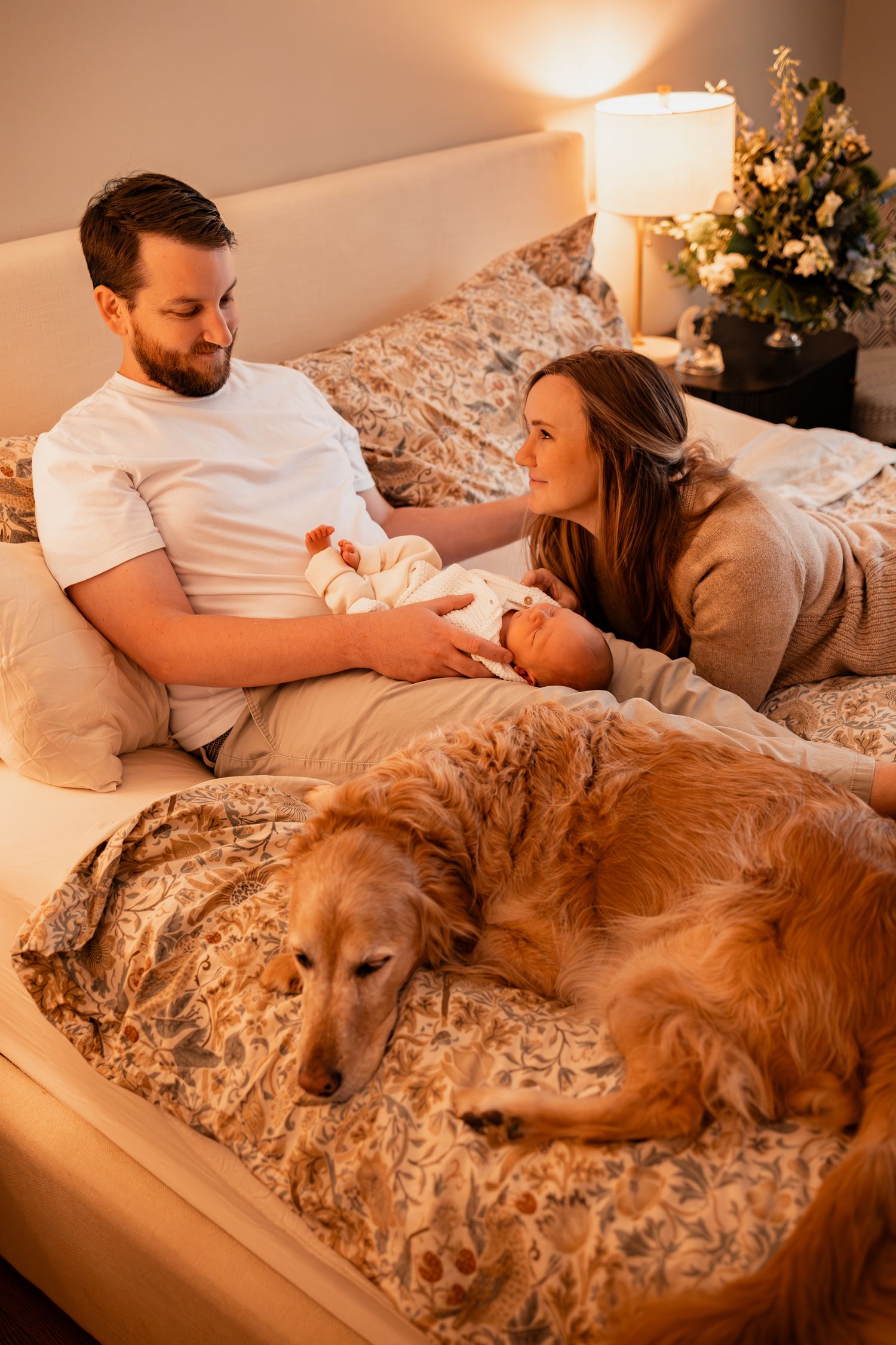 Family lounging on bed with their golden retriever dog in cozy evening light.