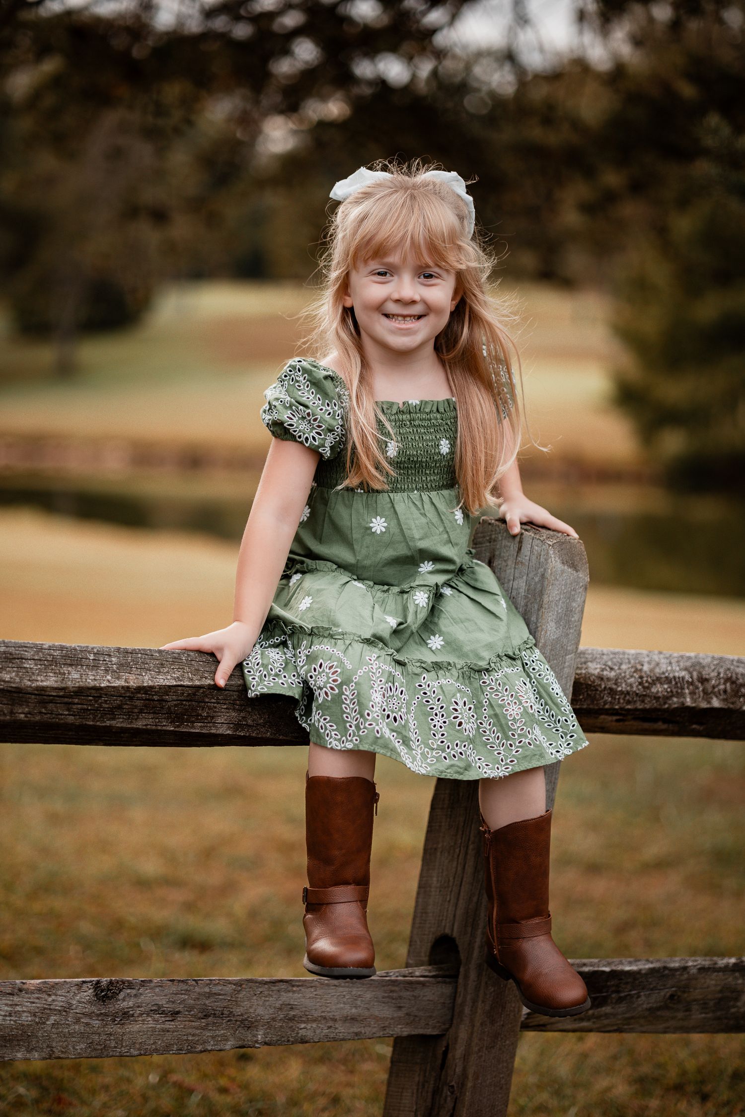 A child in a green eyelet dress and brown boots sits playfully on a rustic wooden fence in an outdoor setting.