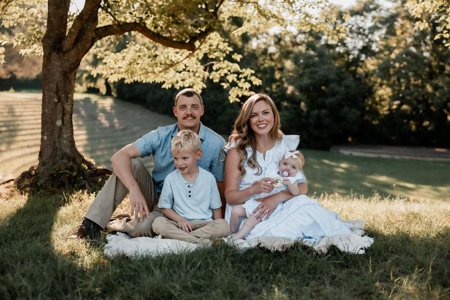 Happy family of four posing outdoors under a tree on a sunny day in a lush green park setting.