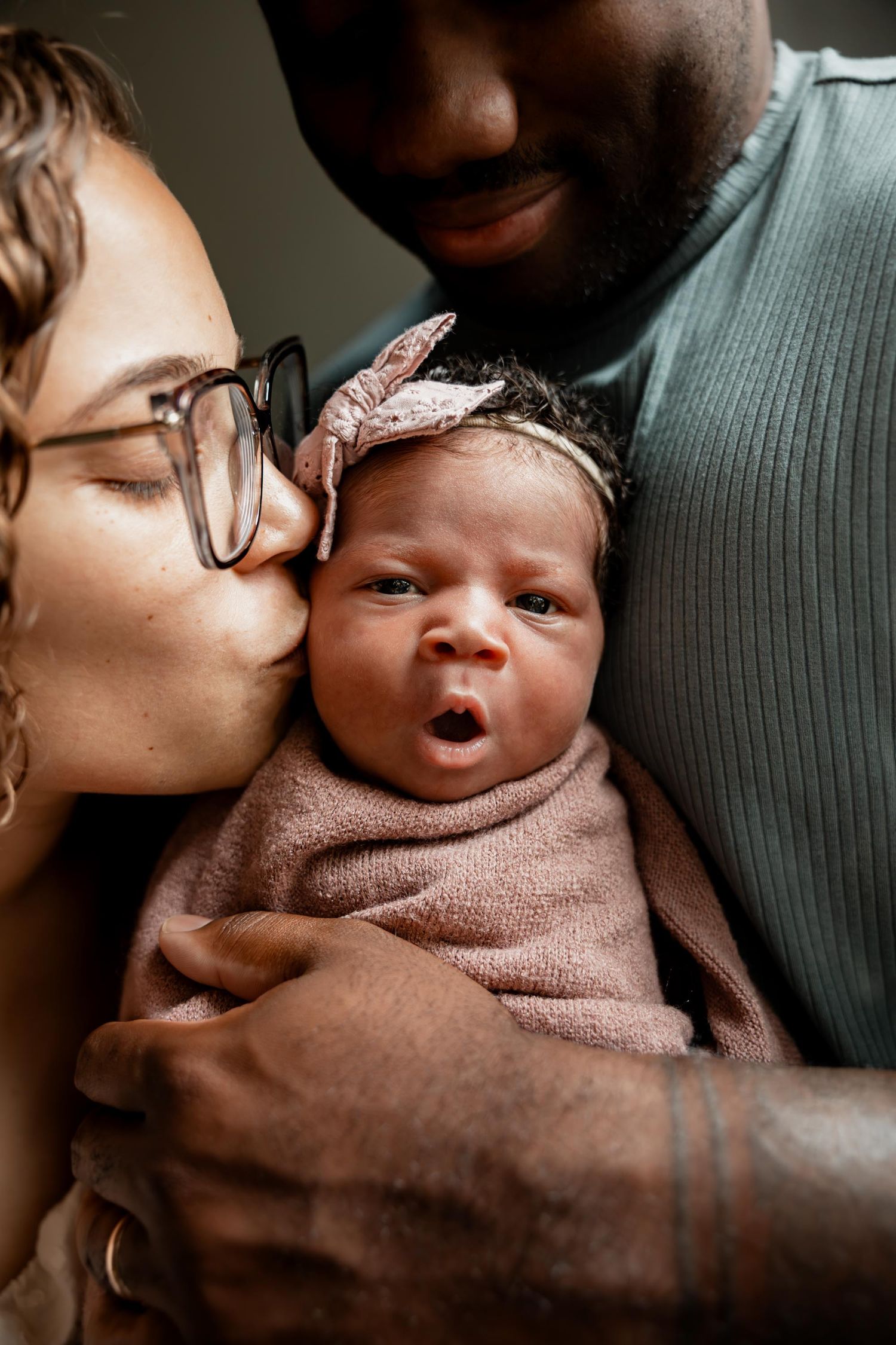 Interracial parents lovingly holding and kissing their swaddled newborn baby girl with bow headband.