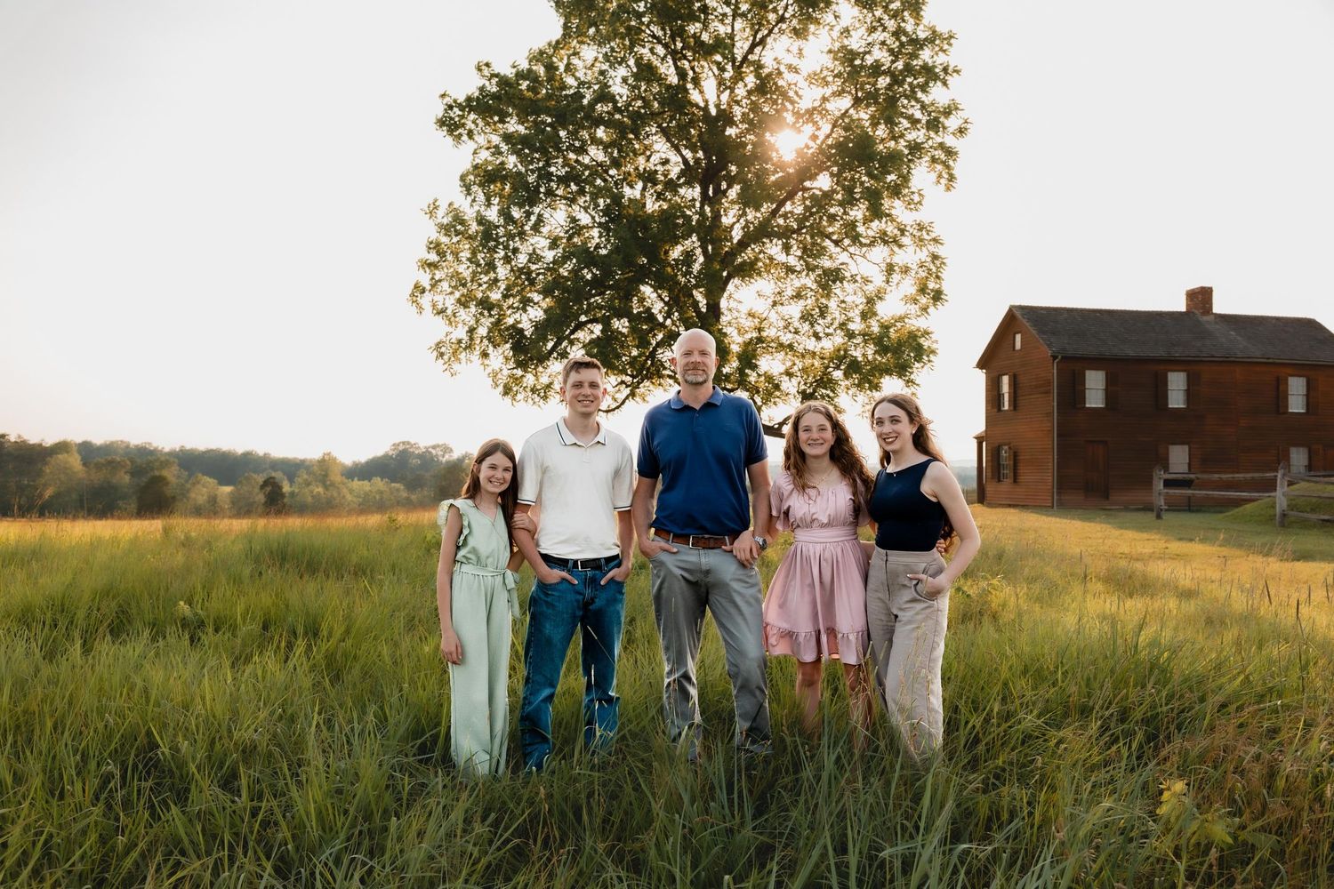 Family of five smiling together in a sunlit green field with a historic brick building and large tree behind them.