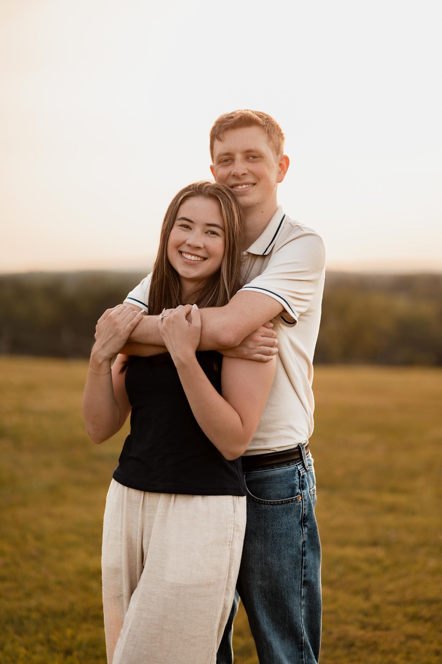 A smiling couple embraces in a green field at sunset, woman in black top and man in white polo shirt.