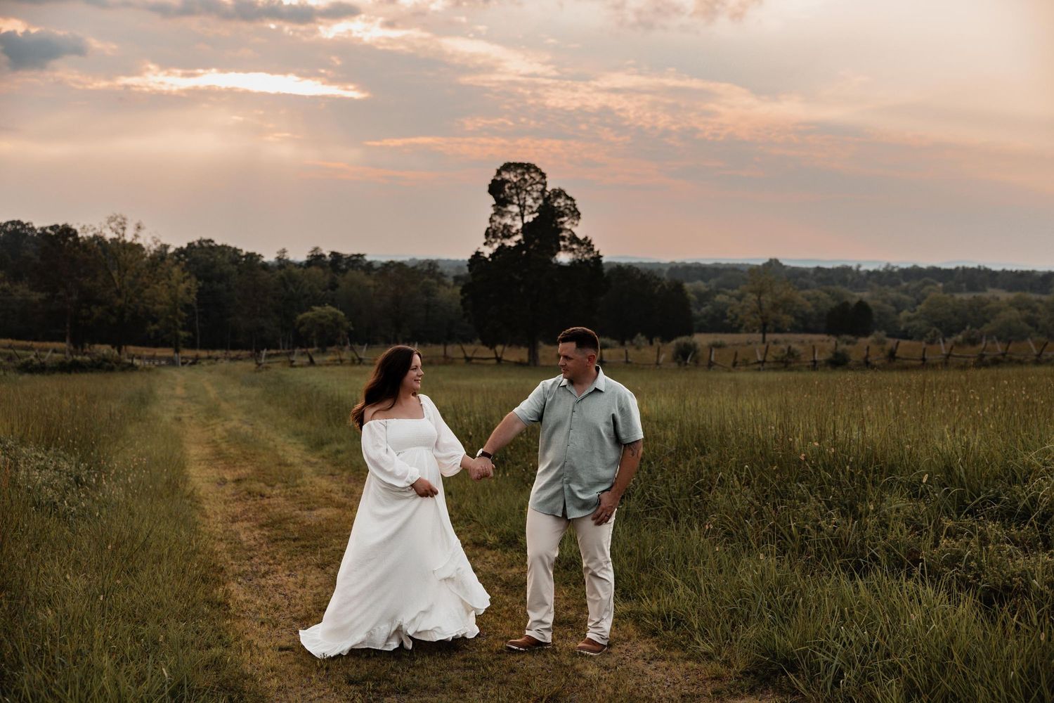 Couple holding hands in a green field at sunset, woman in white gown, man in light blue shirt, romantic outdoor photo.