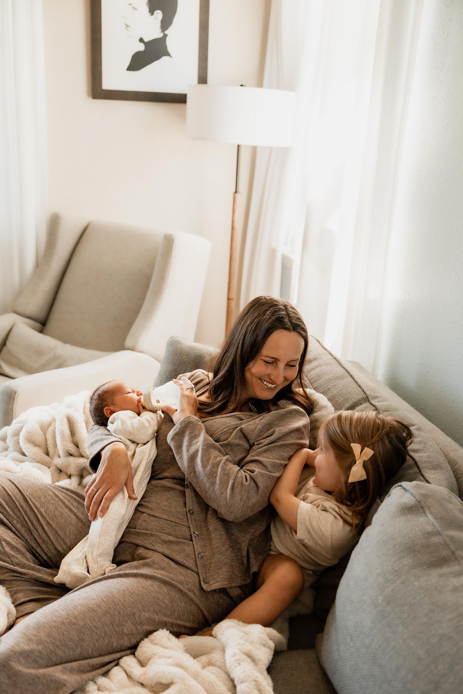 Smiling mother bottle feeds newborn baby while toddler daughter cuddles beside her on sofa.
