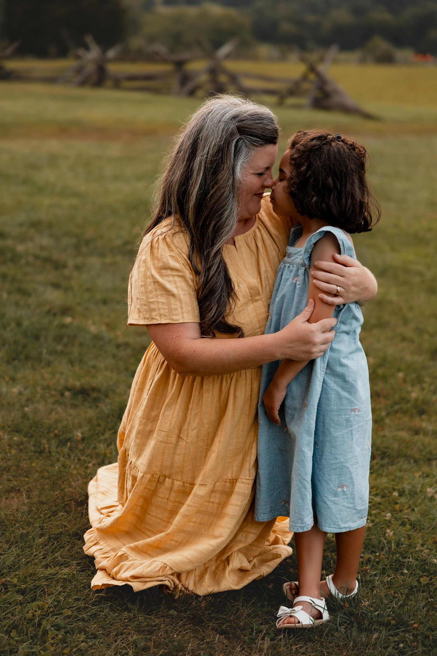 A woman in a yellow maxi dress crouches to embrace a young girl in a blue outfit in a green field.