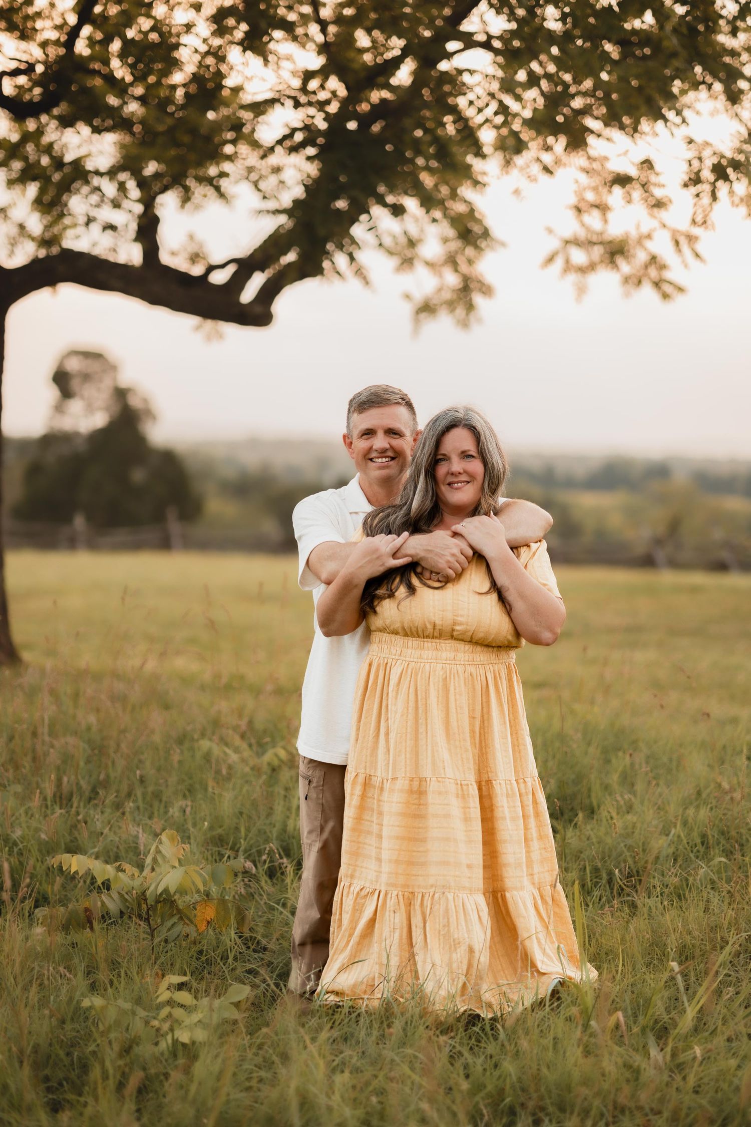 A smiling couple embraces in a sunlit green meadow beneath a large tree at golden hour.