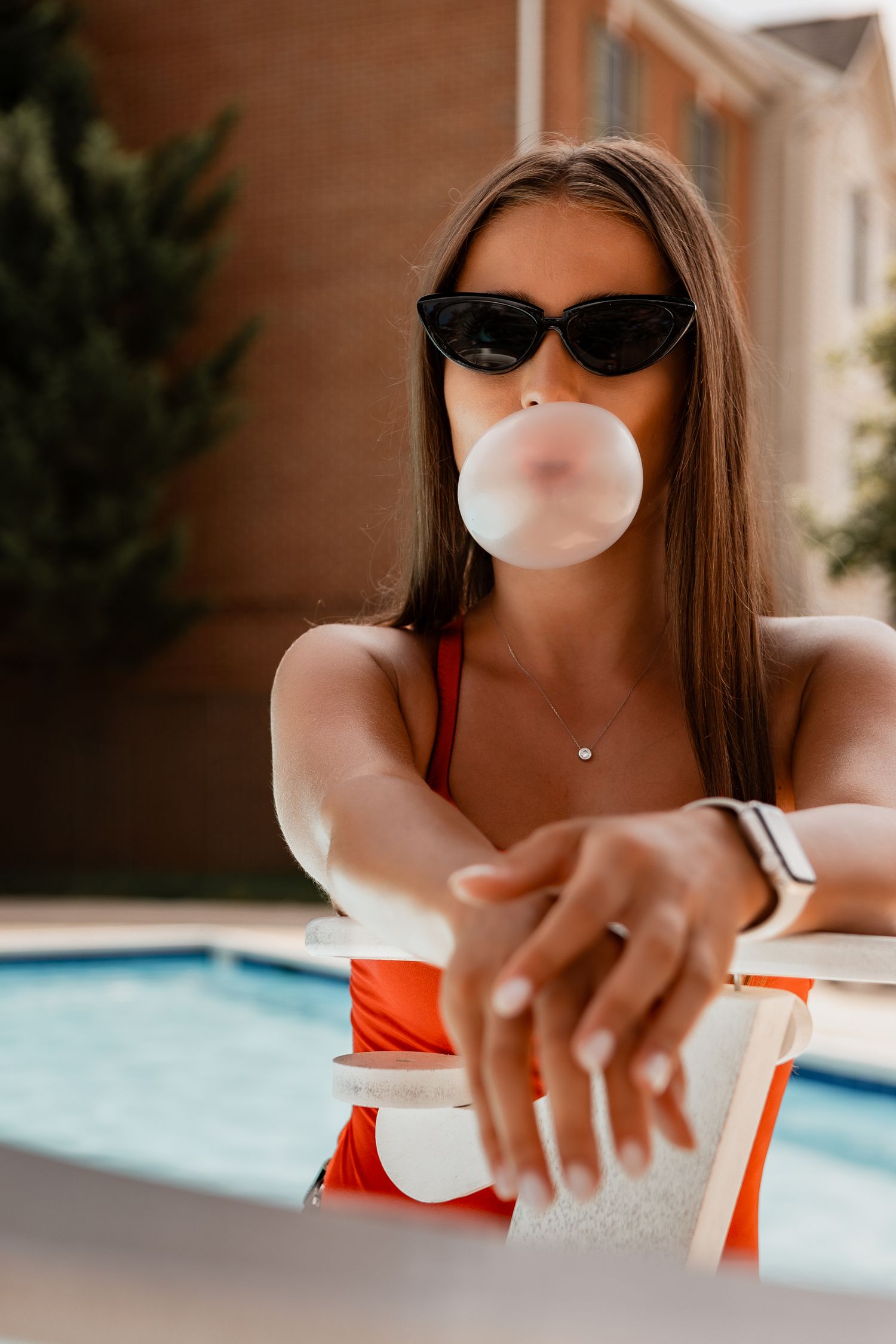 Person in red swimsuit by pool blowing bubble gum and wearing sunglasses.