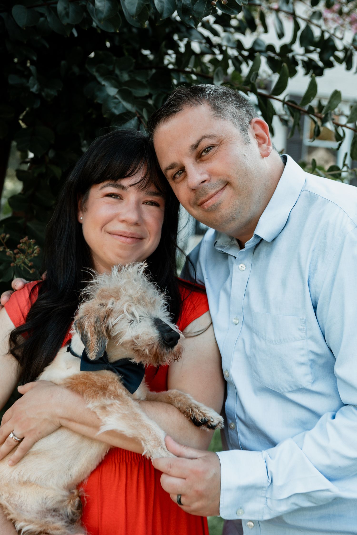 A couple in coral and light blue shirts poses together with their small cream-colored dog in a series of outdoor photos.