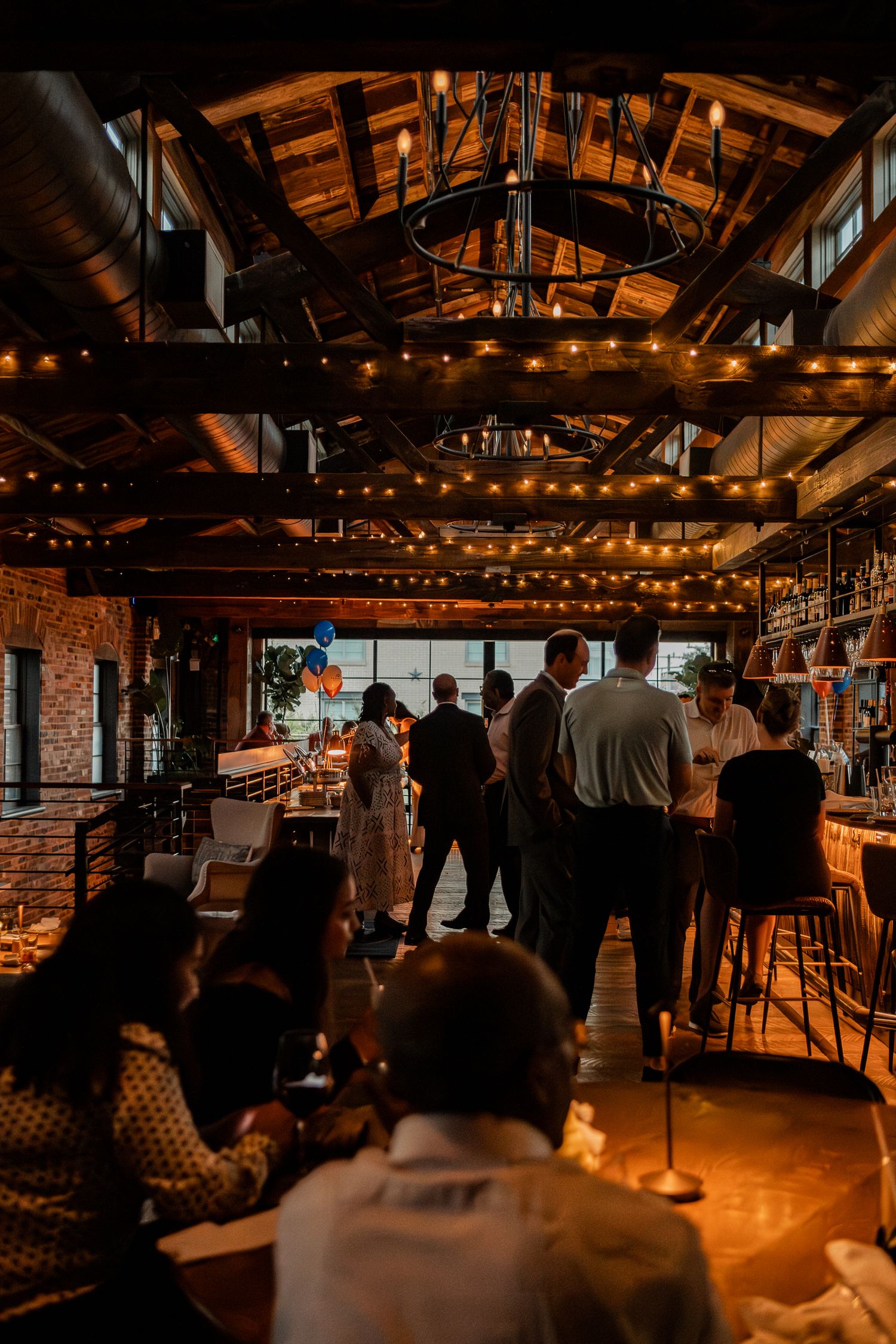 Warm-lit bar interior with exposed wooden beams, string lights, and patrons gathered at counter seats.