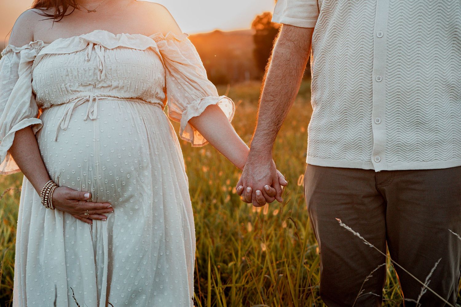 Expectant couple holding hands in golden sunlit meadow during maternity photoshoot.