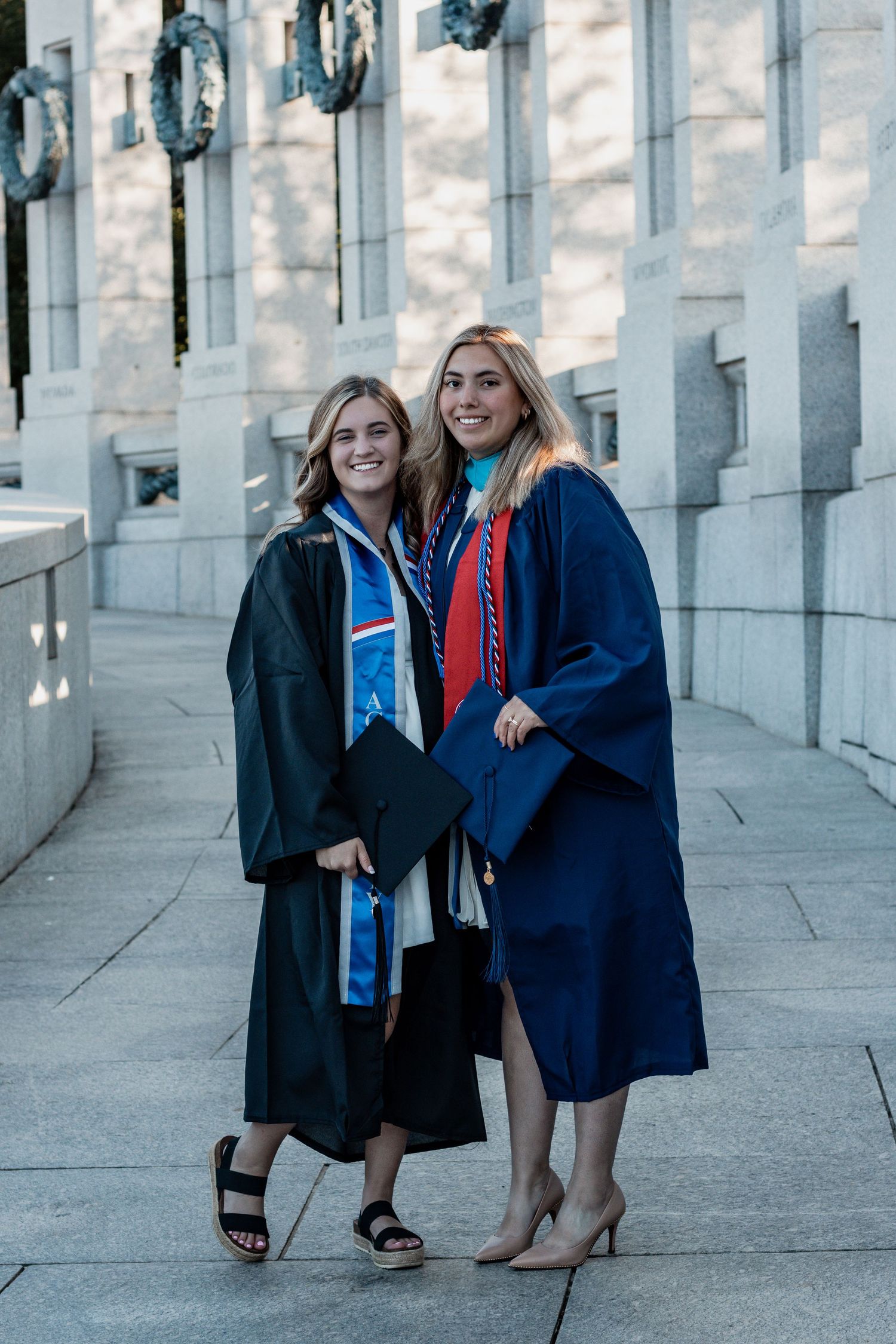 Two graduates posing together in graduation gowns and stoles against white stone columns.