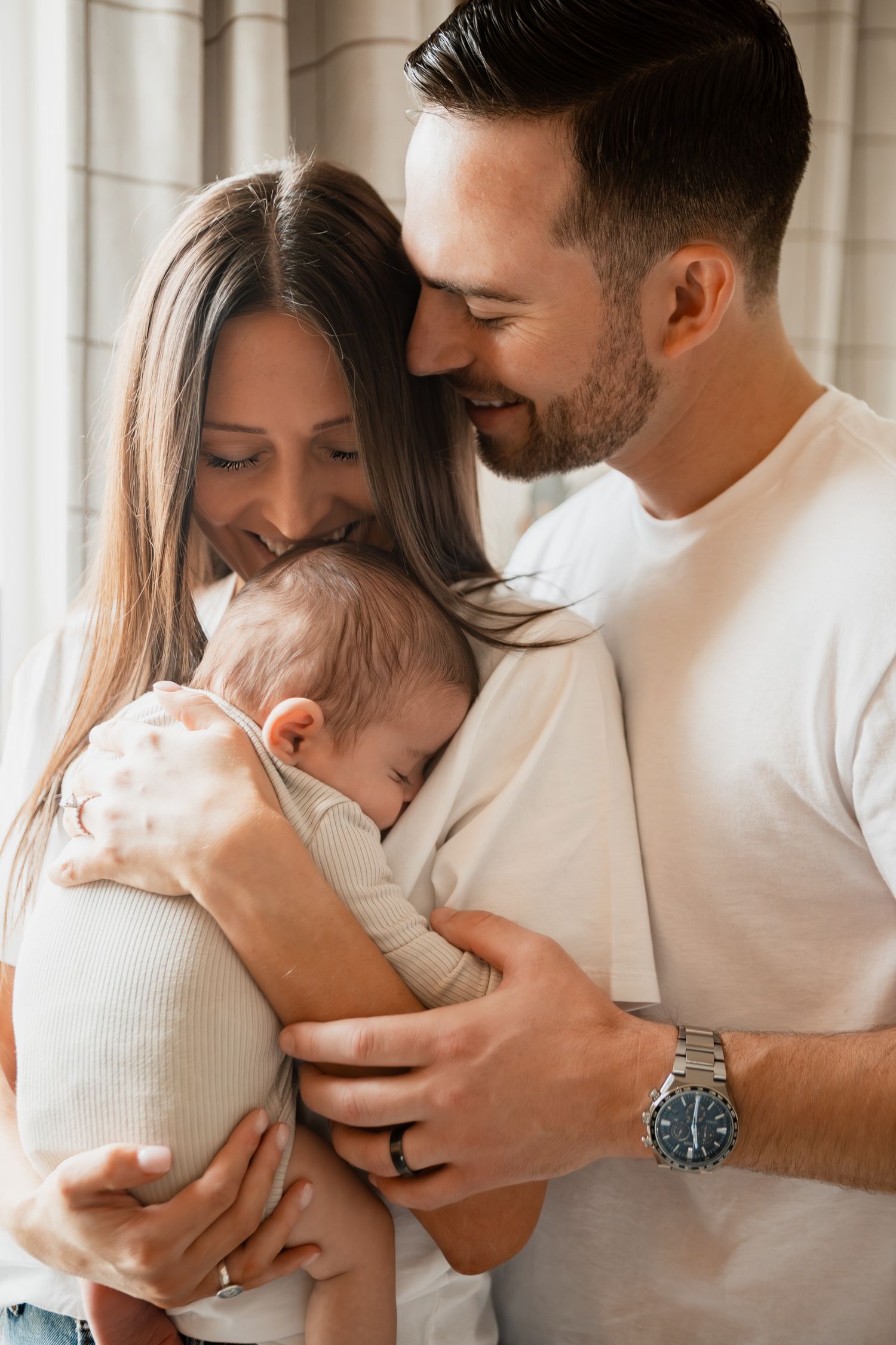 A tender moment between parents embracing their newborn baby in matching white outfits in a collage-style photo.