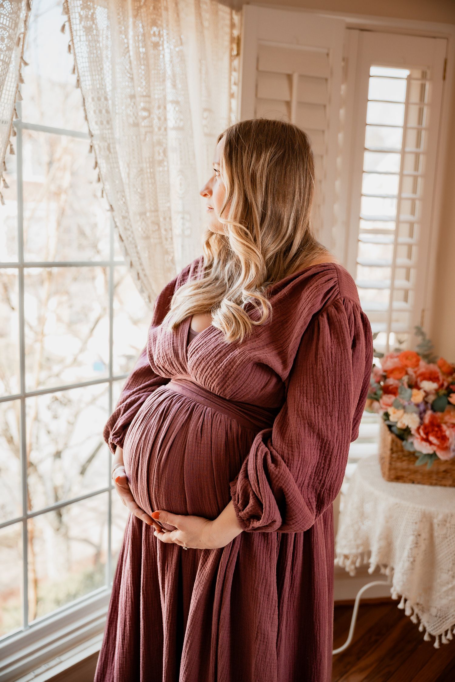 Pregnant woman in dusty rose dress cradling baby bump by window with lace curtains and floral arrangement.