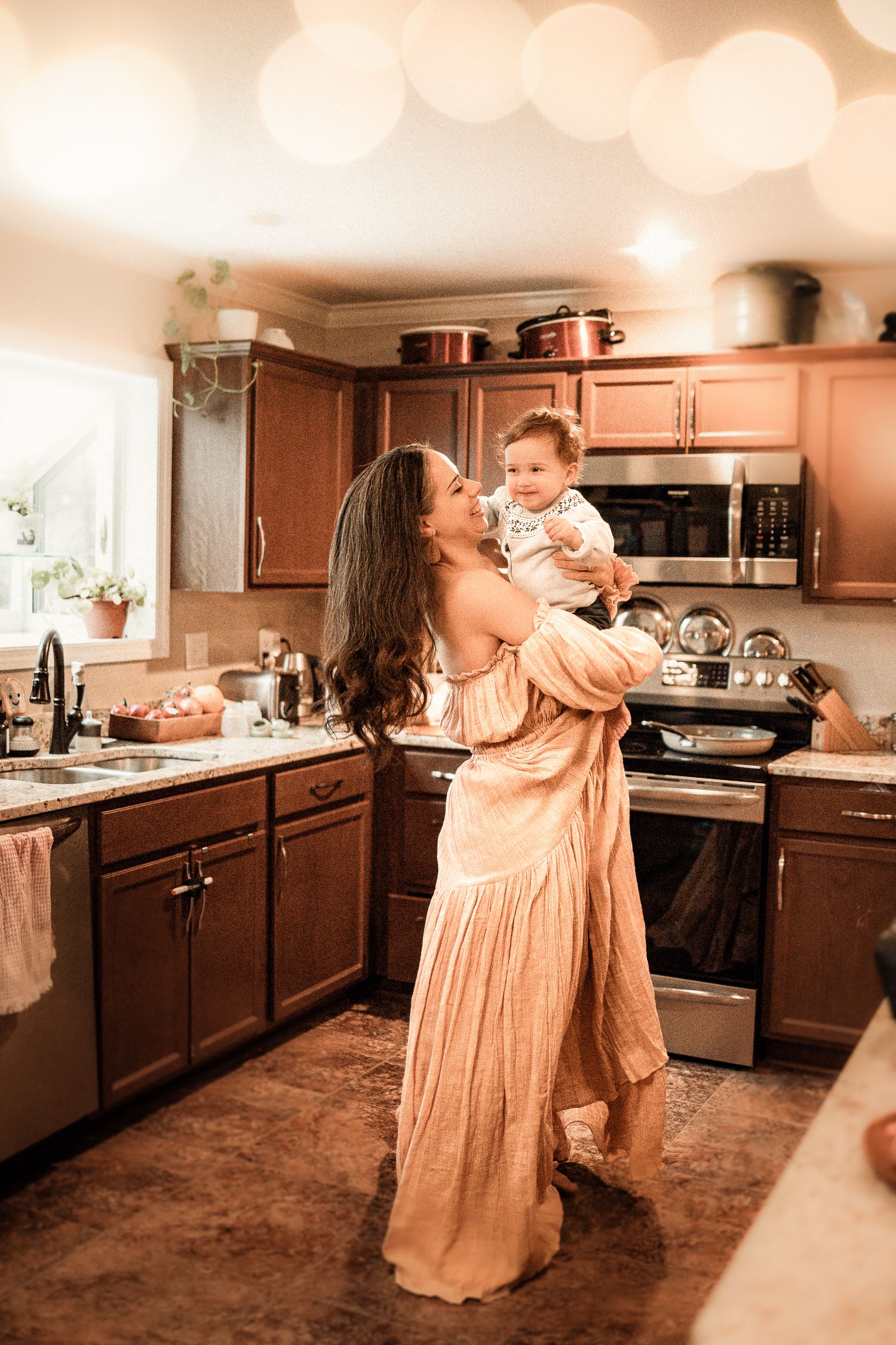 A mother and child dance together in a warm-toned kitchen with dark wood cabinets and vintage style decor.