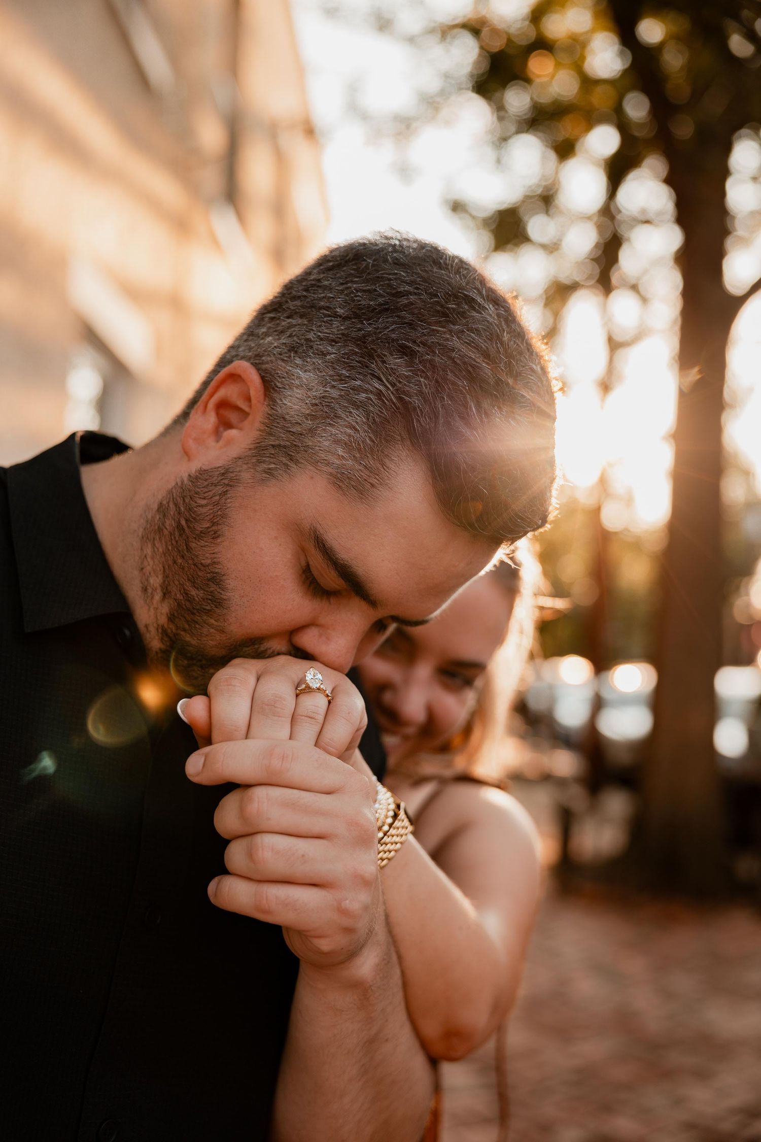 Man kissing woman's hand with engagement ring in golden sunset light outdoors.