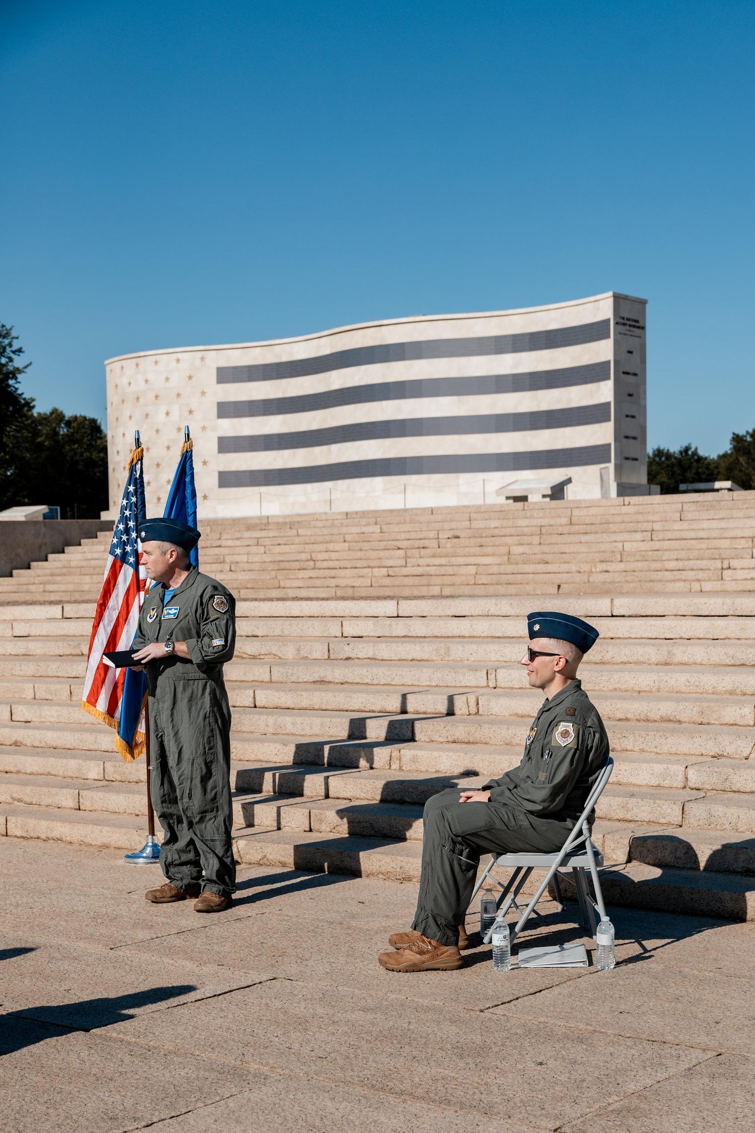 Two military officers in uniform stand near an American flag on marble steps outside a monument on a clear day.