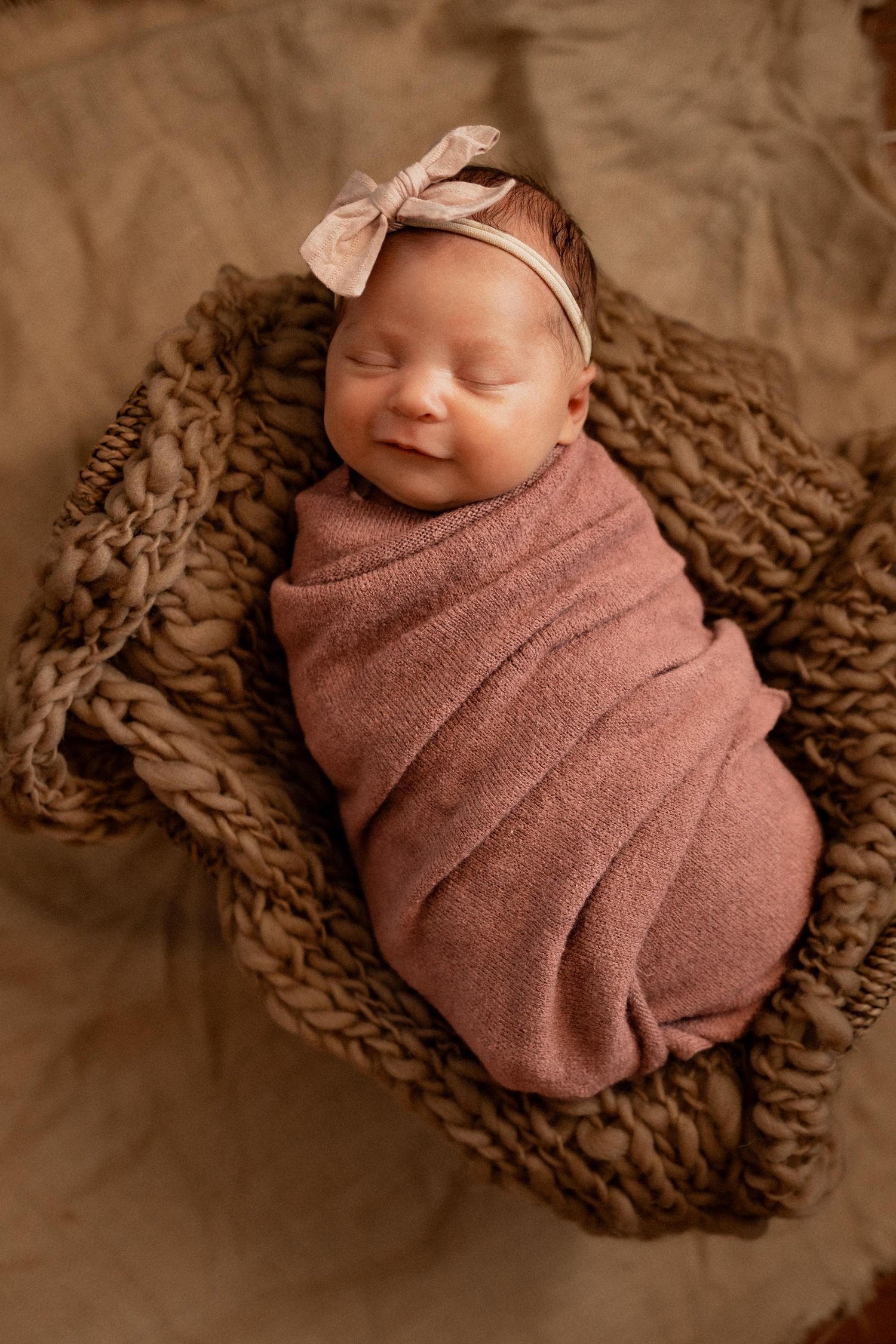 Newborn baby girl swaddled in pink wrap with bow headband, sleeping peacefully in a woven basket.