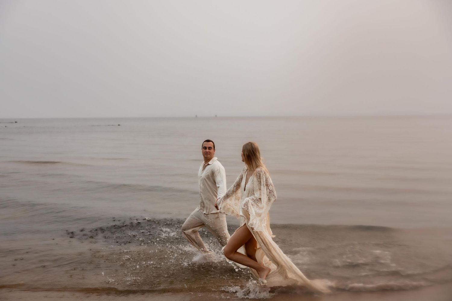 Couple in white wedding attire running through ocean waves on a misty beach shore.