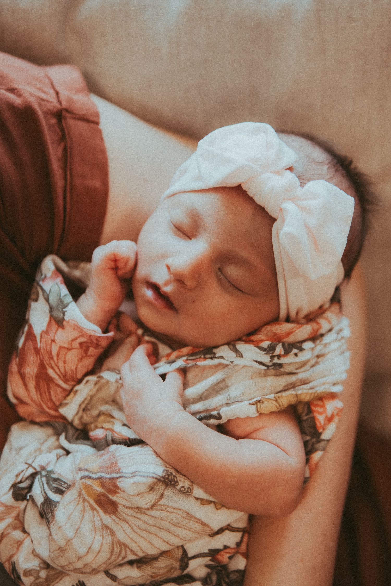 A sleeping baby wearing a floral dress and white bow headband rests peacefully in a warm-toned photograph.