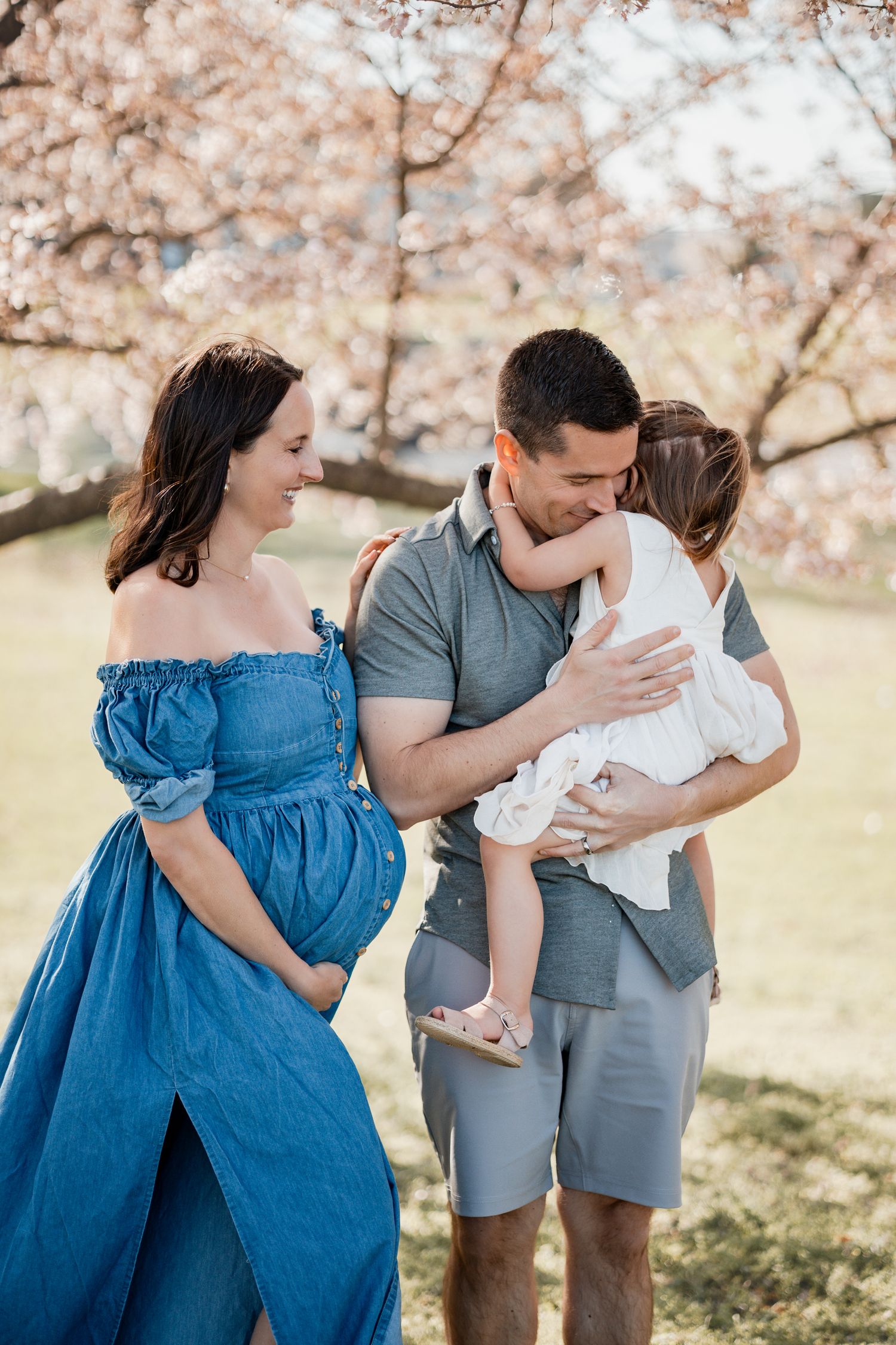 Pregnant woman in blue dress with husband holding toddler under cherry blossom trees during spring family photo session.