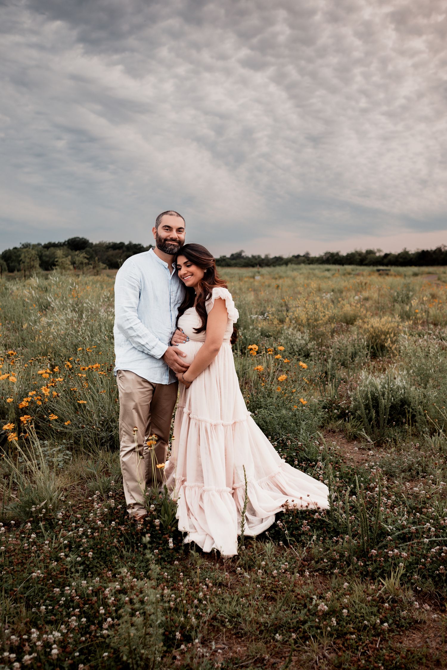 A romantic couple embraces in a wildflower meadow at sunset, with the woman wearing a flowing cream-colored dress.