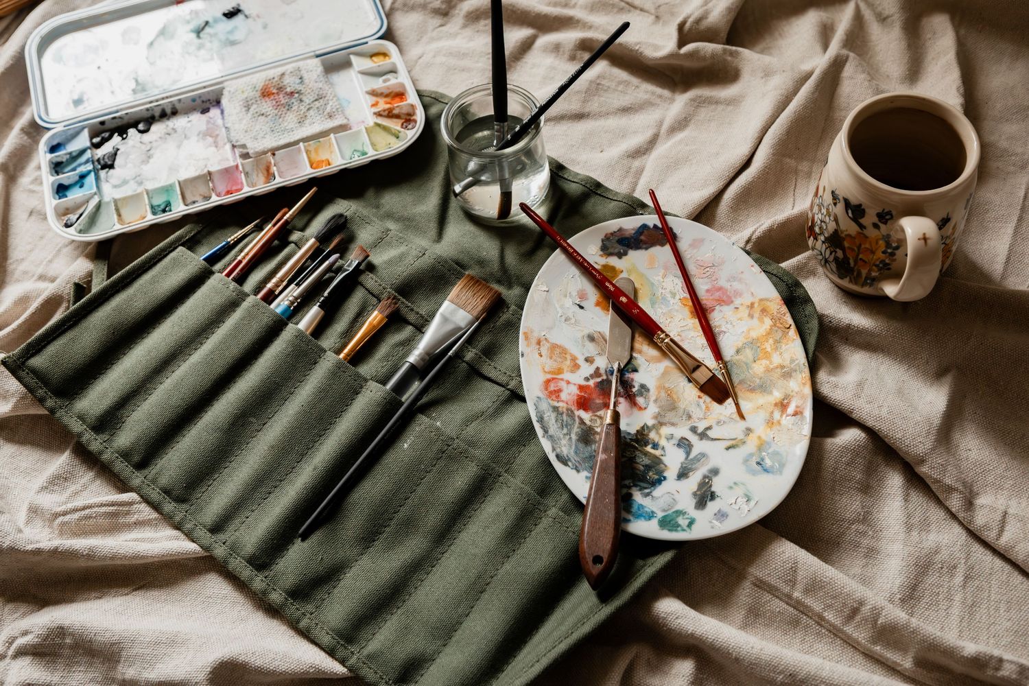 A watercolor art setup with paint brushes in green canvas roll, watercolor palette, and coffee cup on beige fabric.